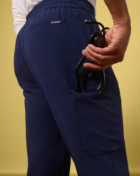 Close-up of the back of a man's Jaanuu Rory Tapered 10-Pocket Scrub Pant in Midnight Navy against a pale yellow wall, showing the JAANUU waistband label, a zip pocket and a cargo pocket holding a black stethoscope. - Men Nav Tile
