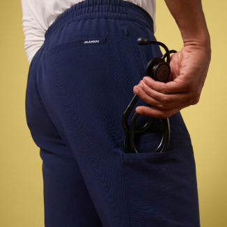 Close-up of the back of a man's Jaanuu Rory Tapered 10-Pocket Scrub Pant in Midnight Navy against a pale yellow wall, showing the JAANUU waistband label, a zip pocket and a cargo pocket holding a black stethoscope. - Men Nav Tile