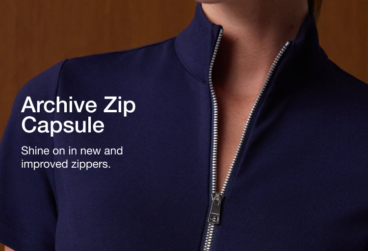 Close-up of a Jaanuu UltraSOFT half-zip scrub top in Midnight Navy + Silver Zip, showing the stand collar and the silver zipper pulled partway up, against a warm wood-paneled wall. Text on the image reads: 'Archive Zip Capsule. Shine on in new and improved zippers.'