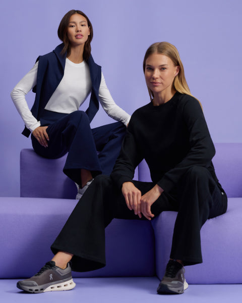Two women in Jaanuu After-Hours loungewear sit on lavender blocks against a lavender wall. One wears the After-Hours Wide Leg Sweatpant in Midnight Navy with the matching After-Hours Crewneck Sweatshirt draped over her shoulders above a white long-sleeve tee, and the other wears the After-Hours Crewneck Sweatshirt and After-Hours Wide Leg Sweatpant in Black. - Women Nav Tile