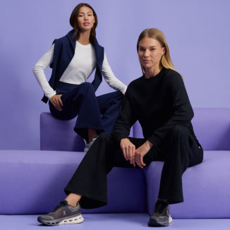 Two women in Jaanuu After-Hours loungewear sit on lavender blocks against a lavender wall. One wears the After-Hours Wide Leg Sweatpant in Midnight Navy with the matching After-Hours Crewneck Sweatshirt draped over her shoulders above a white long-sleeve tee, and the other wears the After-Hours Crewneck Sweatshirt and After-Hours Wide Leg Sweatpant in Black. - Women Nav Tile