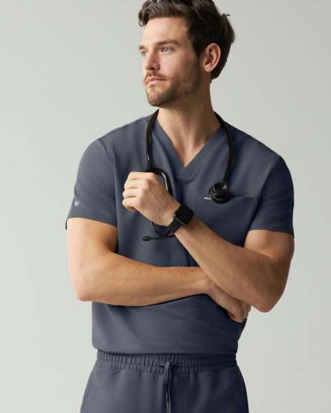 A man in a slate grey Jaanuu V-neck scrub top stands with his arms crossed, a stethoscope around his neck and a smartwatch on his wrist, looking to the side against a light grey wall. - Men Nav Tile