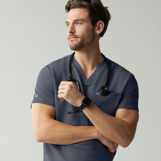 A man in a slate grey Jaanuu V-neck scrub top stands with his arms crossed, a stethoscope around his neck and a smartwatch on his wrist, looking to the side against a light grey wall. - Men Nav Tile