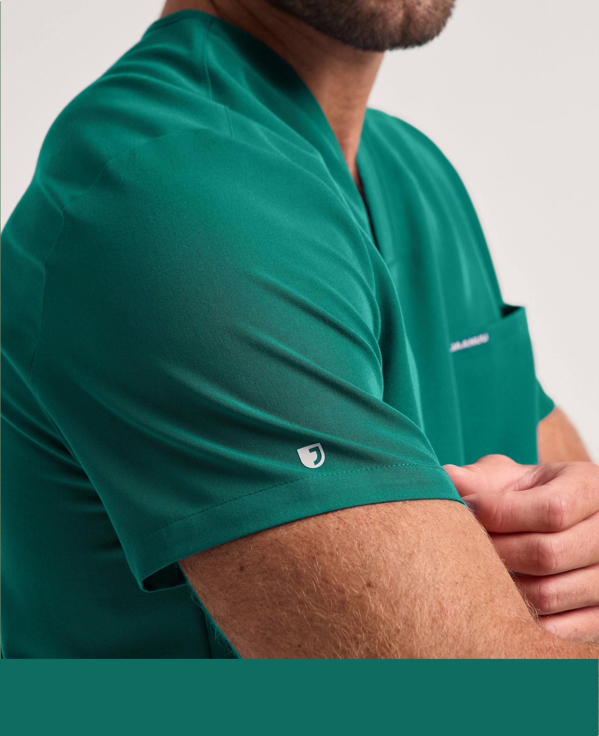 A close-up of a bearded man with his arms crossed in a Hunter Green (dark green) Jaanuu V-neck scrub top with a chest pocket and a logo on the sleeve, against a light grey background.