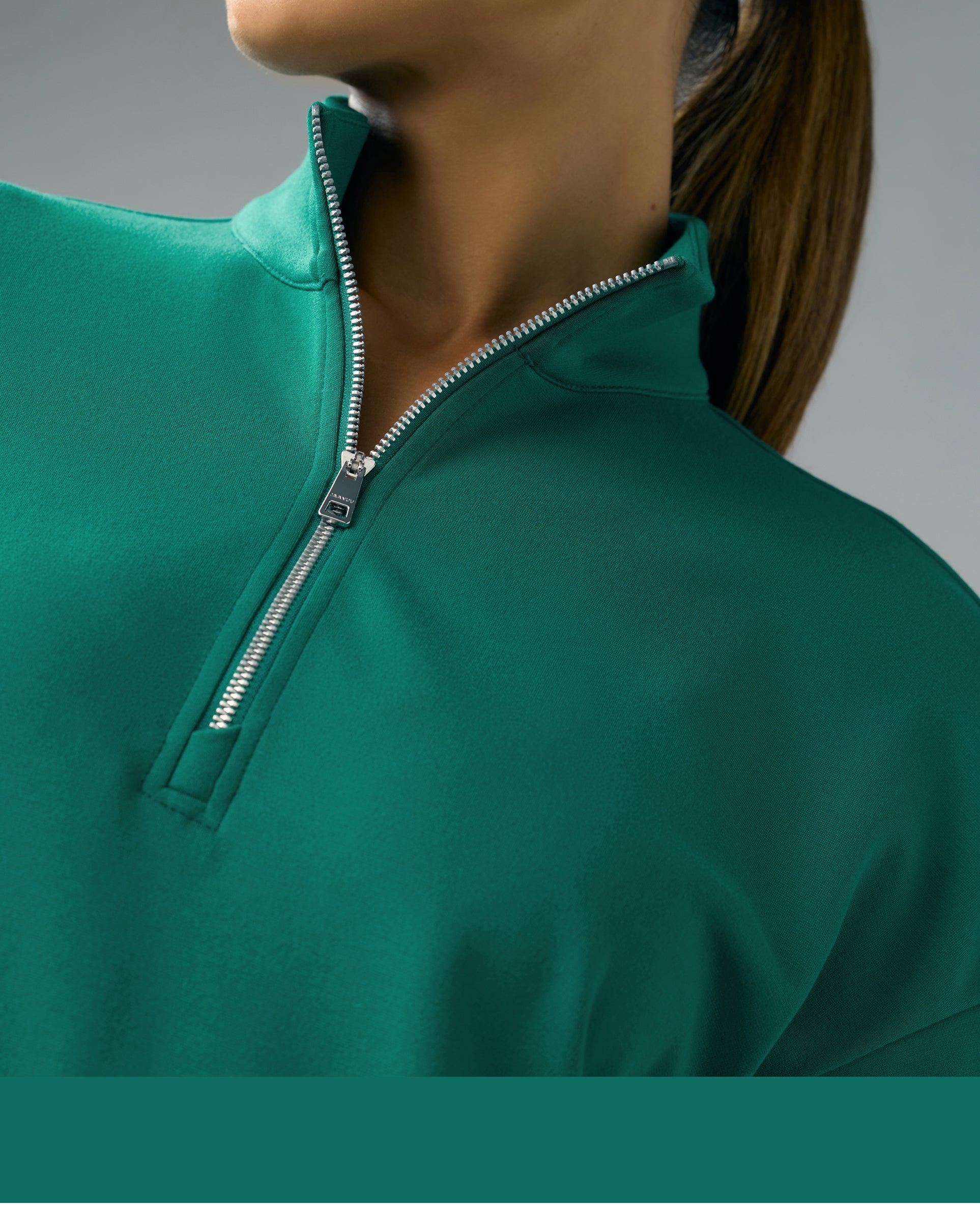 A close-up of a woman with a ponytail in a Hunter Green (dark green) Jaanuu half-zip scrub top with a silver zipper, against a grey background.