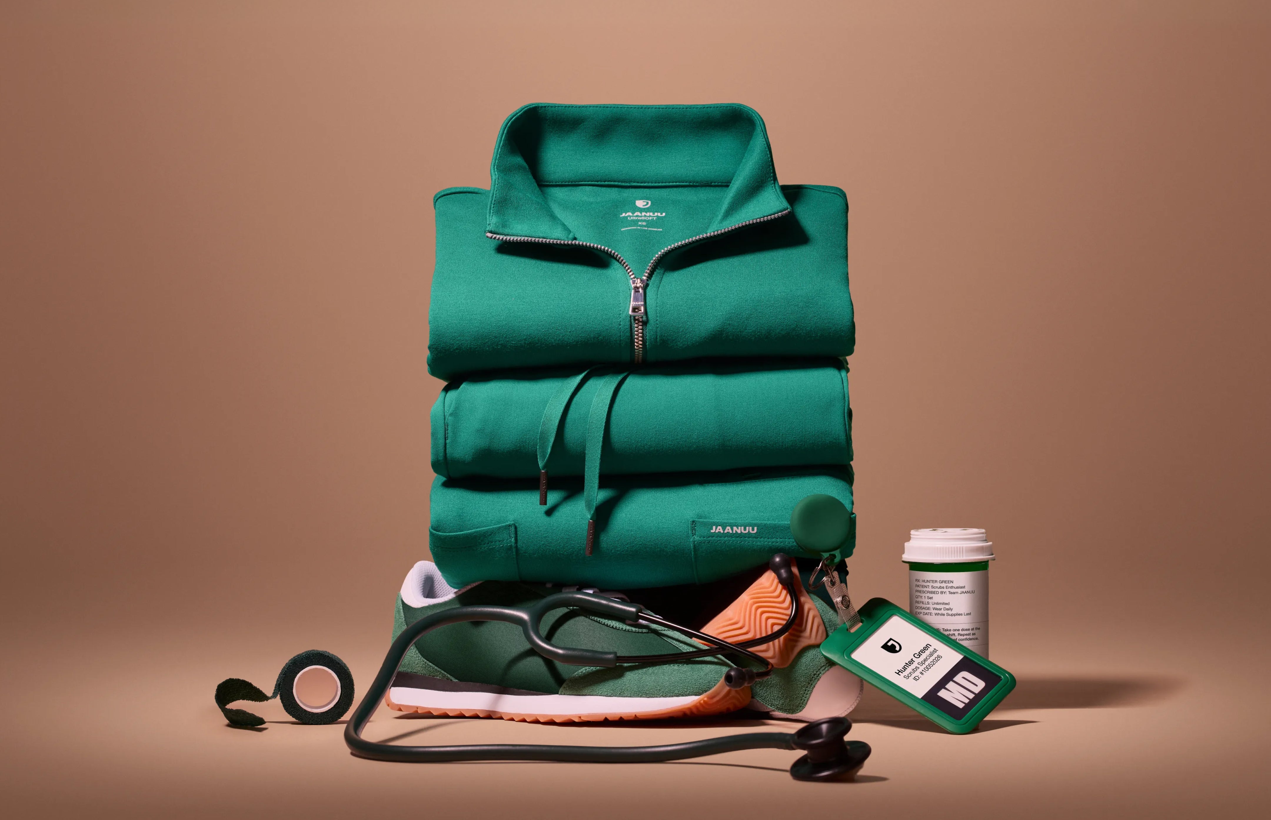 A stack of folded Hunter Green (dark green) Jaanuu scrubs topped by a half-zip scrub top, with a green sneaker, a stethoscope, a roll of green tape, a green ID badge reading 'Hunter Green' and a prescription-style pill bottle at its base, against a tan backdrop.
