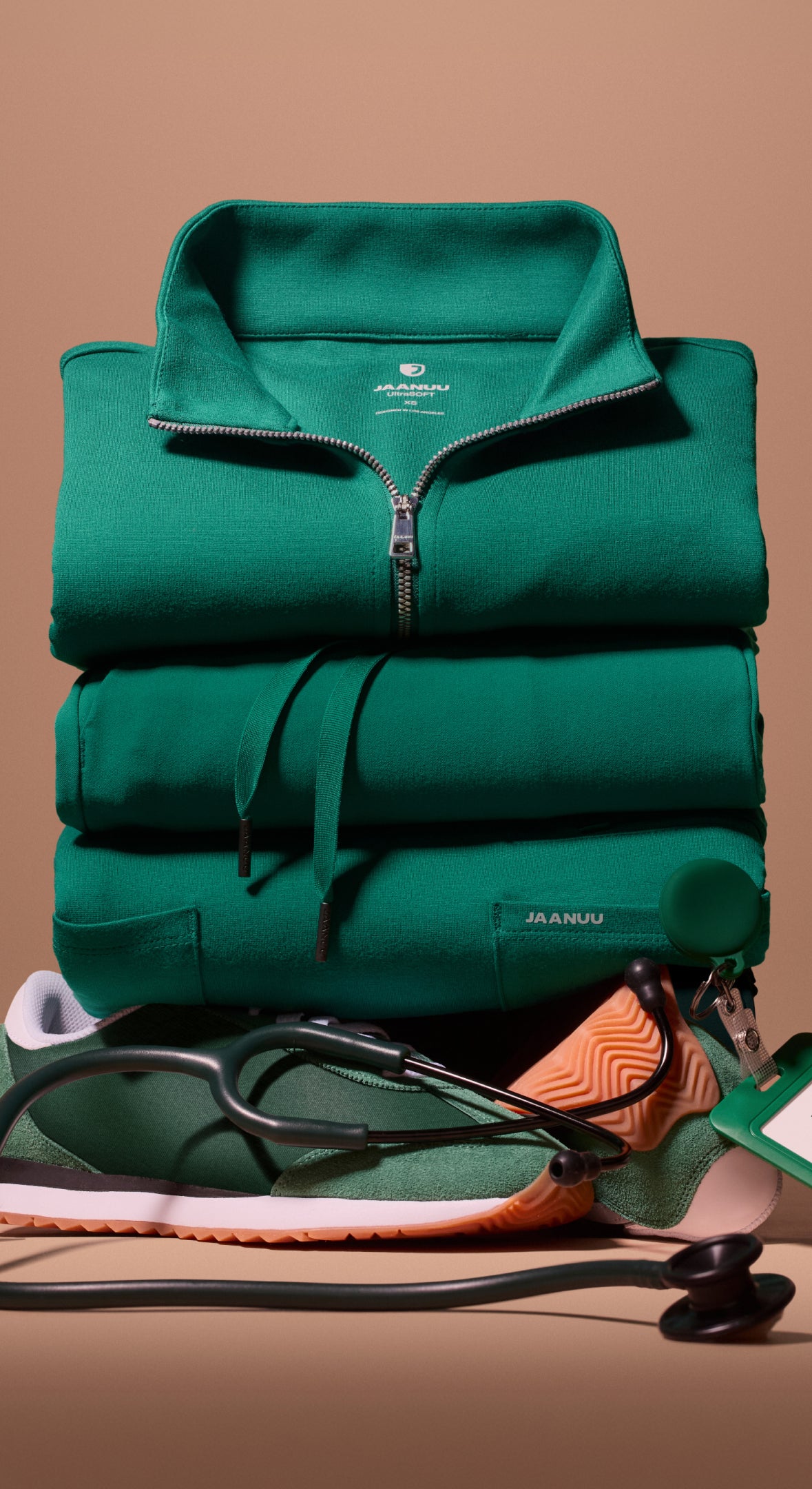 A stack of folded Hunter Green (dark green) Jaanuu scrubs topped by a half-zip scrub top, with a green sneaker, a stethoscope and a green ID badge at its base, against a tan backdrop.