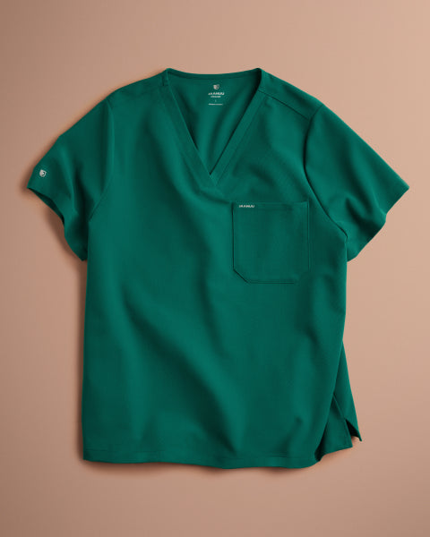 A Hunter Green (dark green) Jaanuu V-neck scrub top with a chest pocket and a logo on the sleeve, laid flat on a blush pink background. - Men Nav Tile