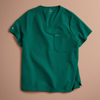 A Hunter Green (dark green) Jaanuu V-neck scrub top with a chest pocket and a logo on the sleeve, laid flat on a blush pink background. - Men Nav Tile