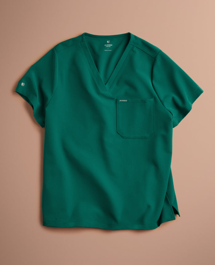 A Hunter Green (dark green) Jaanuu V-neck scrub top with a chest pocket and a logo on the sleeve, laid flat on a blush pink background.