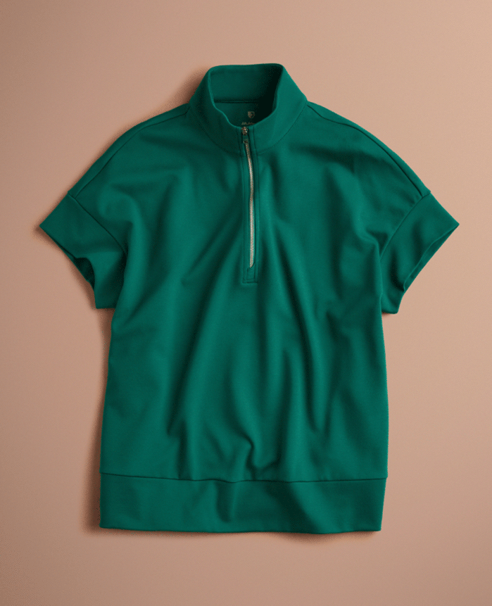 Animated flat lay that alternates between a Hunter Green (dark green) Jaanuu short-sleeve half-zip scrub top and matching flare-leg scrub pants with a zippered leg pocket, on a blush pink background.