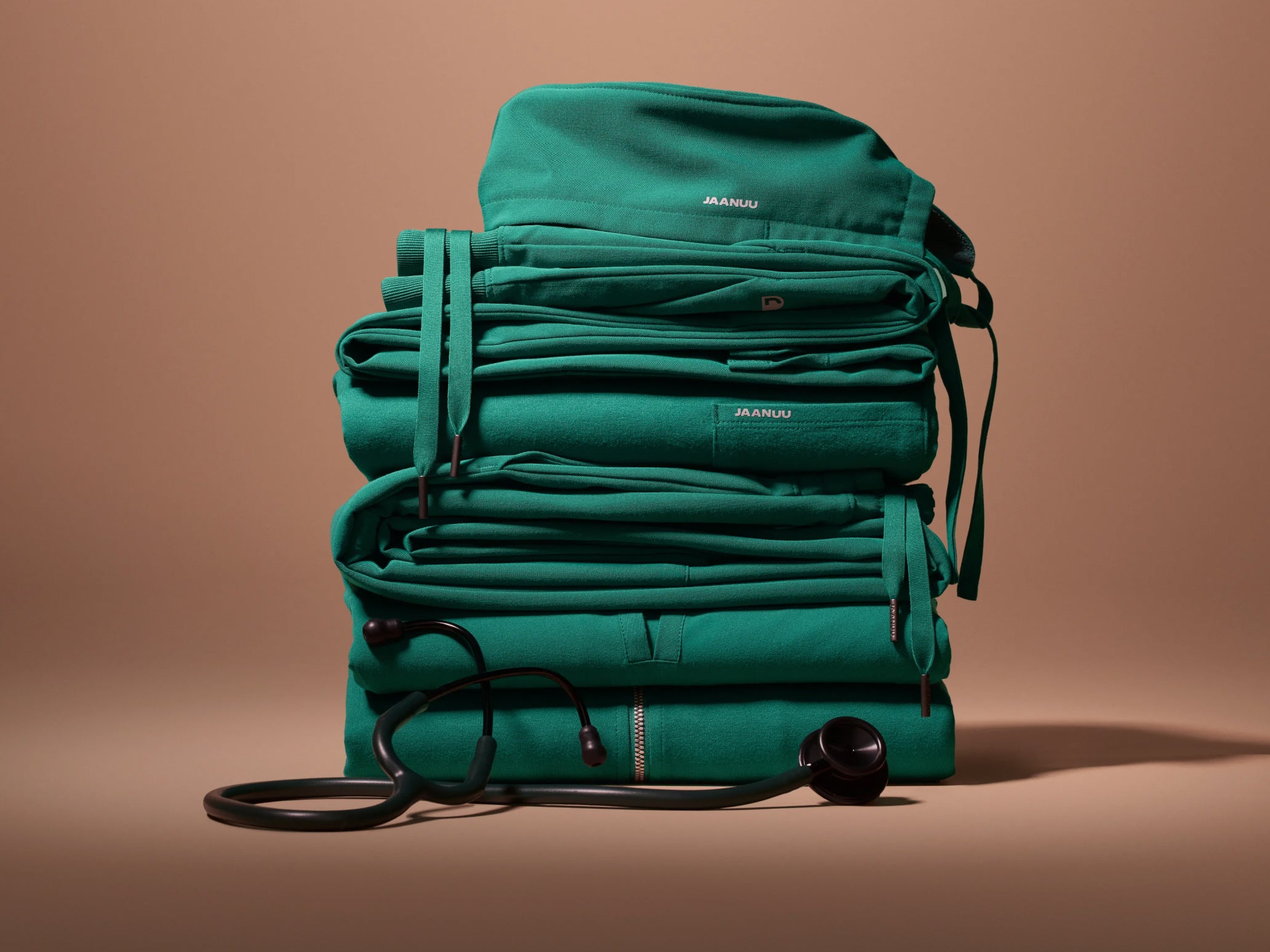 A tall stack of folded Hunter Green (dark green) Jaanuu scrubs with drawstrings hanging down the sides and a stethoscope draped across the base, against a tan backdrop.