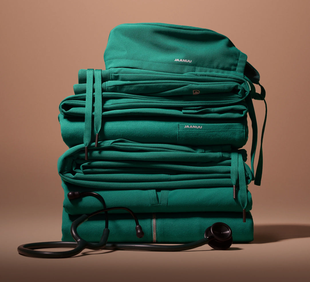 A tall stack of folded Hunter Green (dark green) Jaanuu scrubs with drawstrings hanging down the sides and a stethoscope draped across the base, against a tan backdrop.