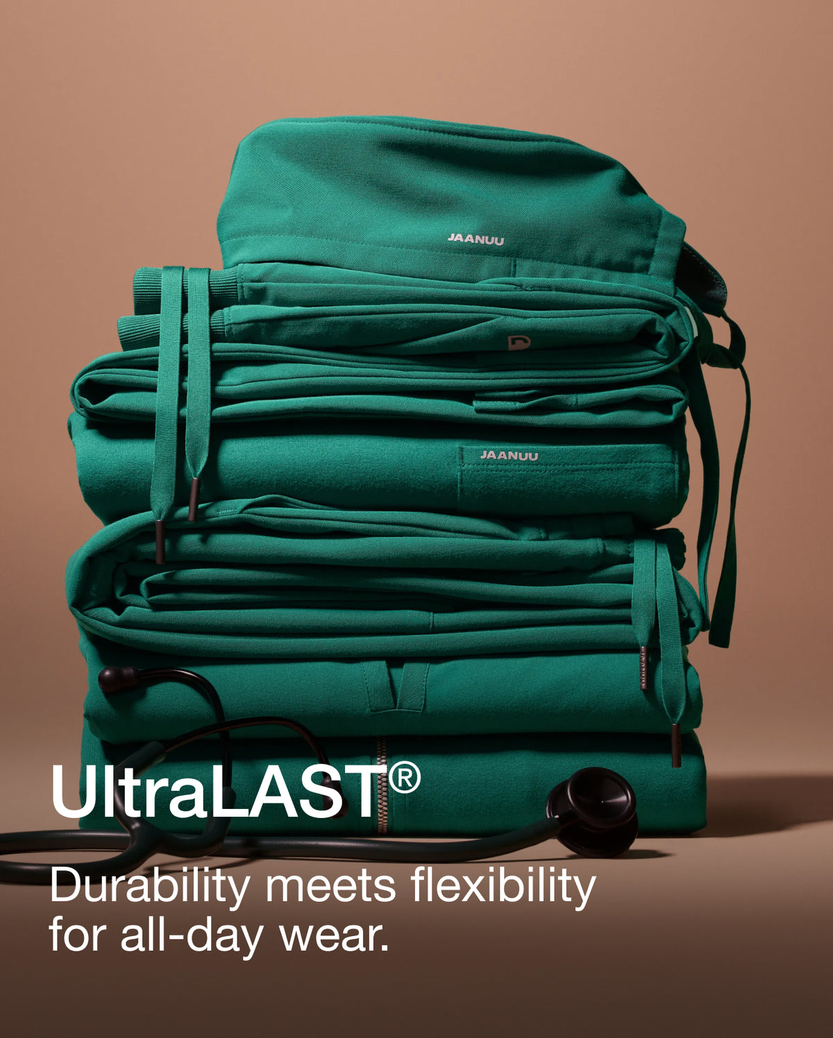 A tall stack of folded Hunter Green (dark green) Jaanuu scrubs with drawstrings hanging down the sides and a stethoscope at the base, against a tan backdrop. Text on the image reads: 'UltraLAST®. Durability meets flexibility for all-day wear.'