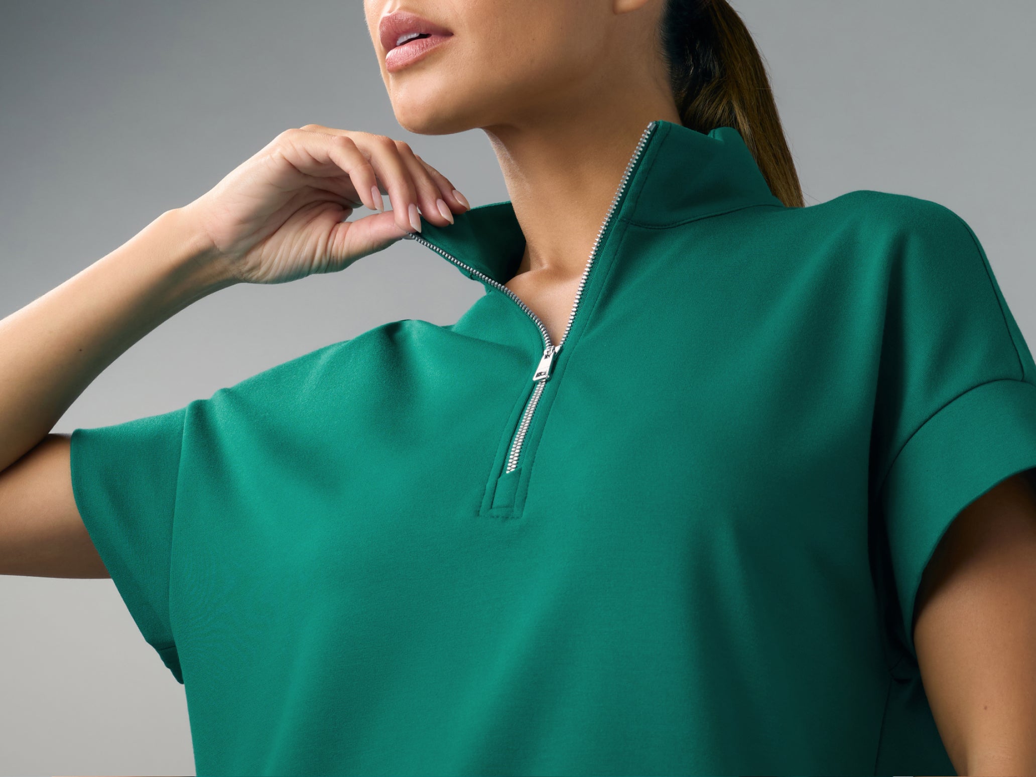 A close-up of a woman with a ponytail in a Hunter Green (dark green) Jaanuu short-sleeve half-zip scrub top, holding the open collar with one hand, against a grey background.
