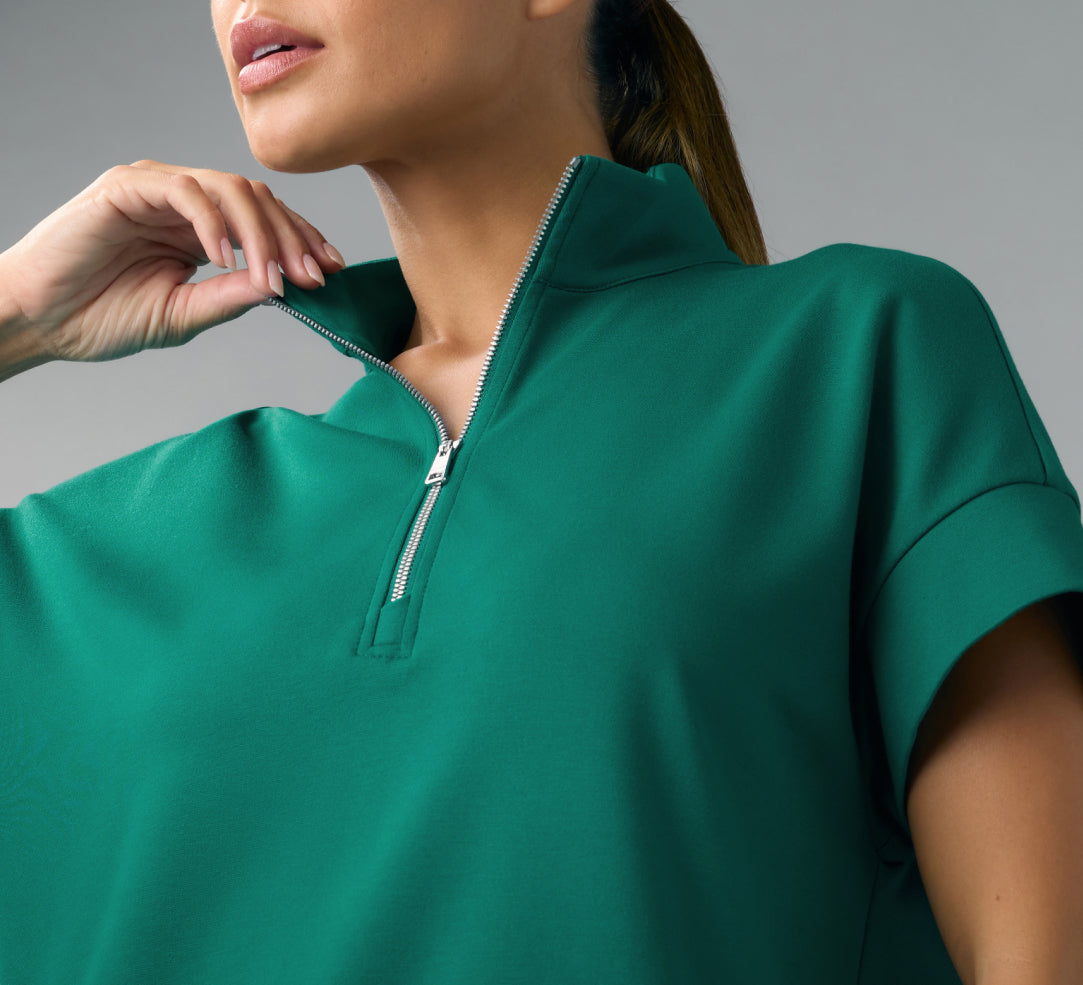 A close-up of a woman with a ponytail in a Hunter Green (dark green) Jaanuu short-sleeve half-zip scrub top, holding the open collar with one hand, against a grey background.