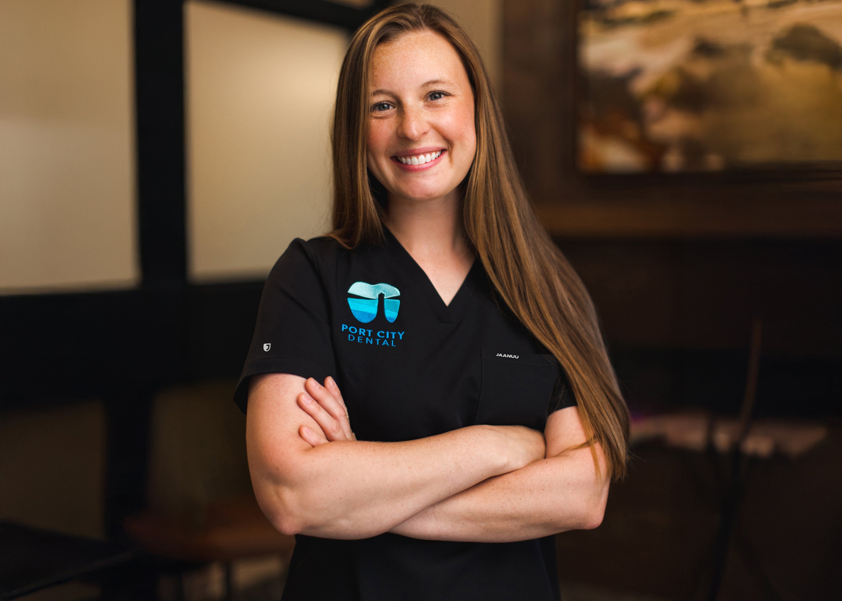 Smiling woman with long hair and folded arms in a black Jaanuu scrub top embroidered with the Port City Dental logo