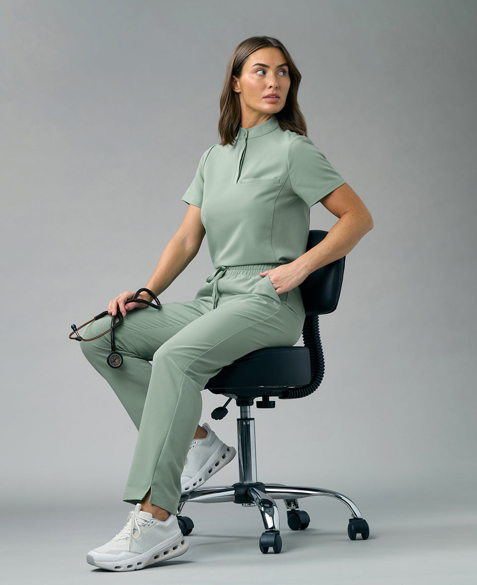 Woman in a pale green Jaanuu scrub top and pants sitting on a black rolling stool with a stethoscope in her hand