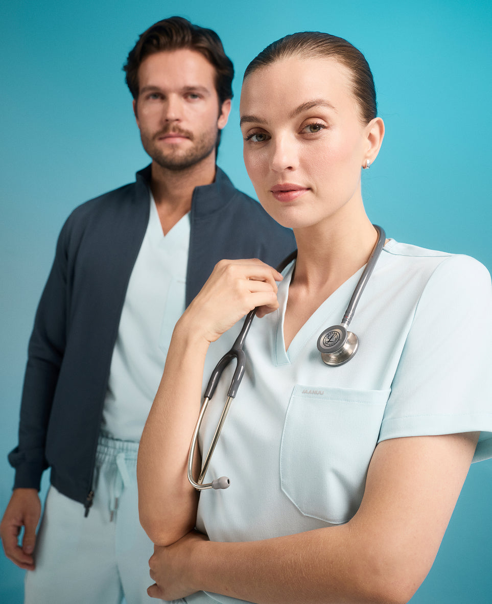 Woman in a light blue Jaanuu scrub top with a stethoscope around her neck, and behind her a man in a dark zip jacket over light blue scrubs, against a turquoise backdrop