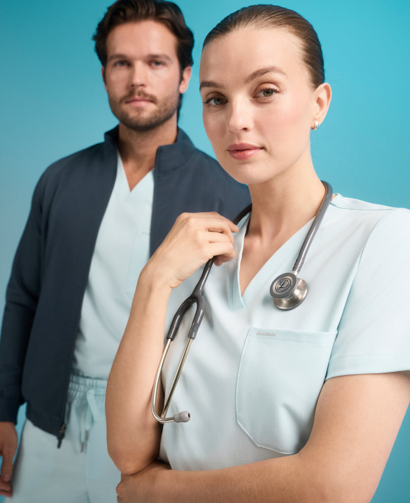 Woman in a light blue Jaanuu scrub top with a stethoscope around her neck, and behind her a man in a dark zip jacket over light blue scrubs, against a turquoise backdrop