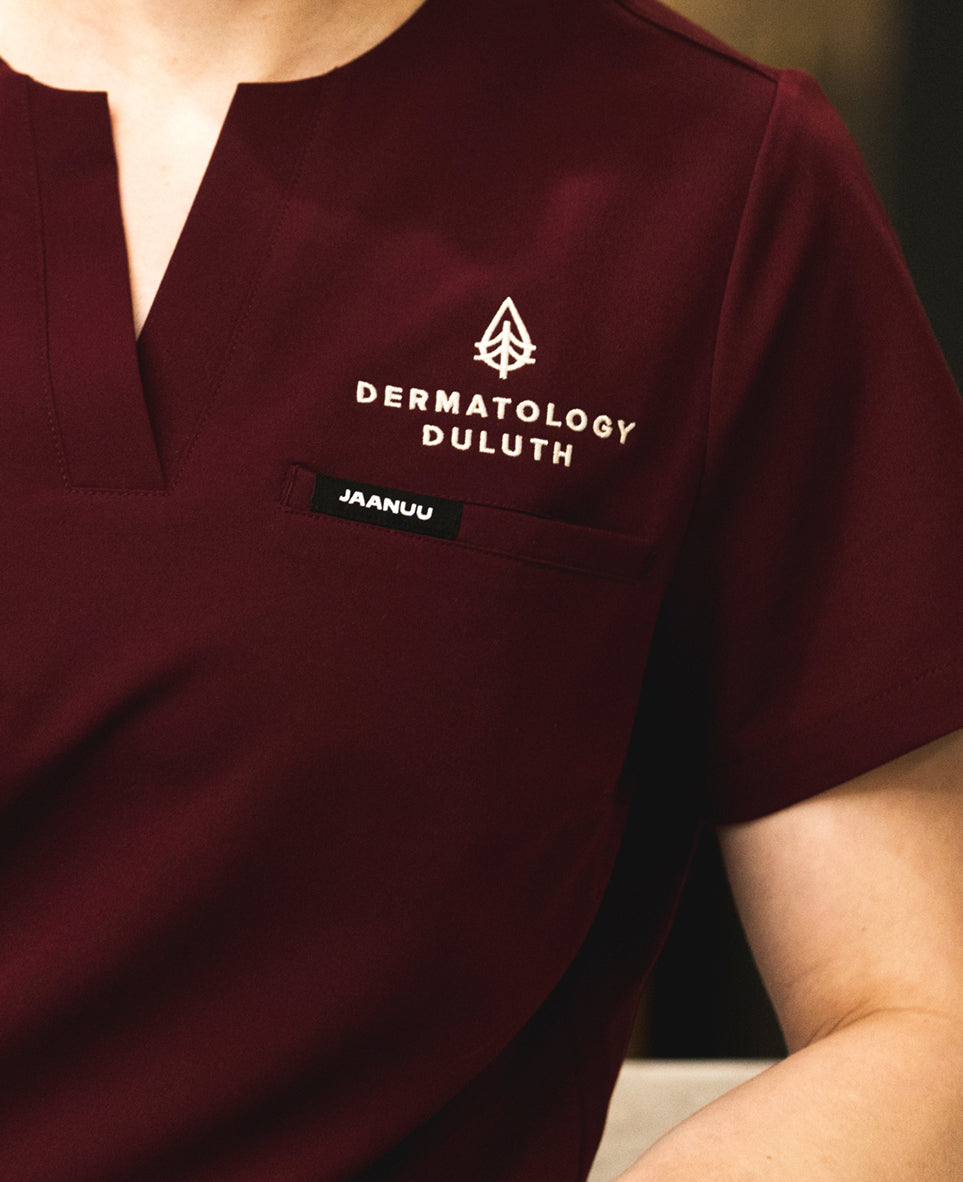 Detail of a dark red Jaanuu scrub top embroidered with the Dermatology Duluth logo above the chest pocket