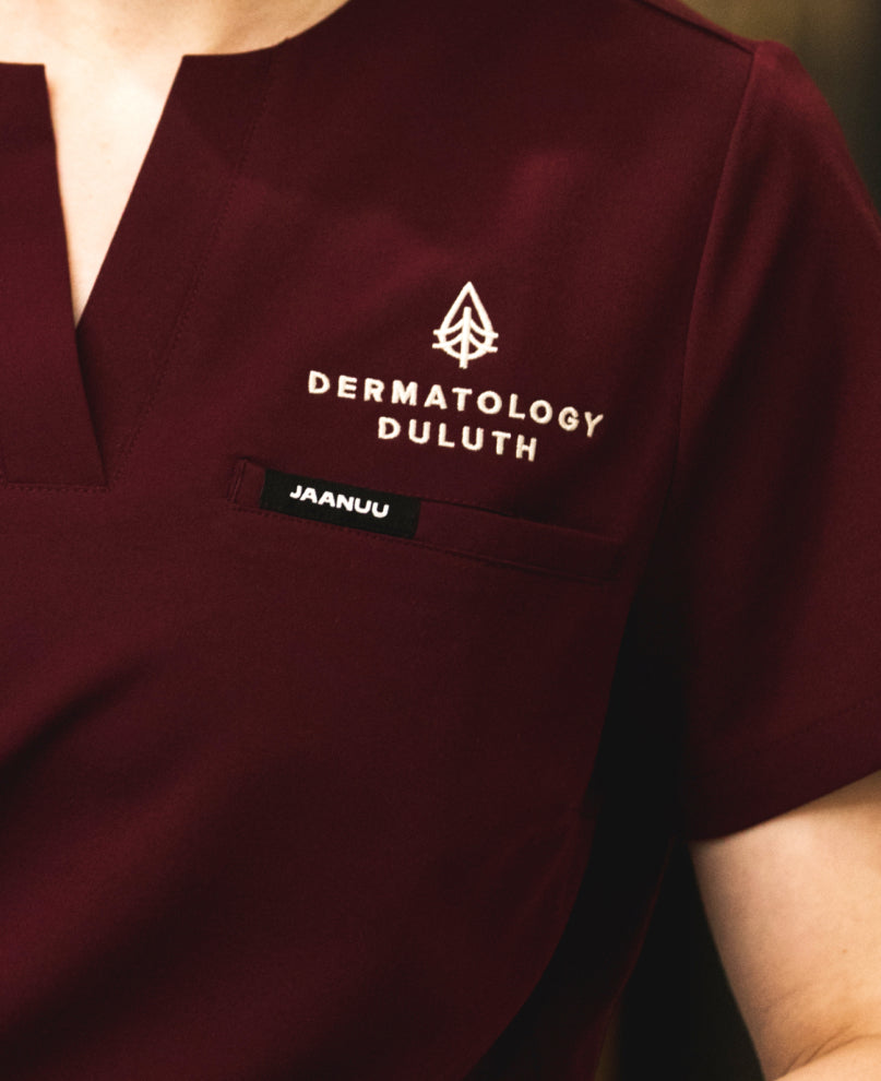 Detail of a dark red Jaanuu scrub top embroidered with the Dermatology Duluth logo above the chest pocket