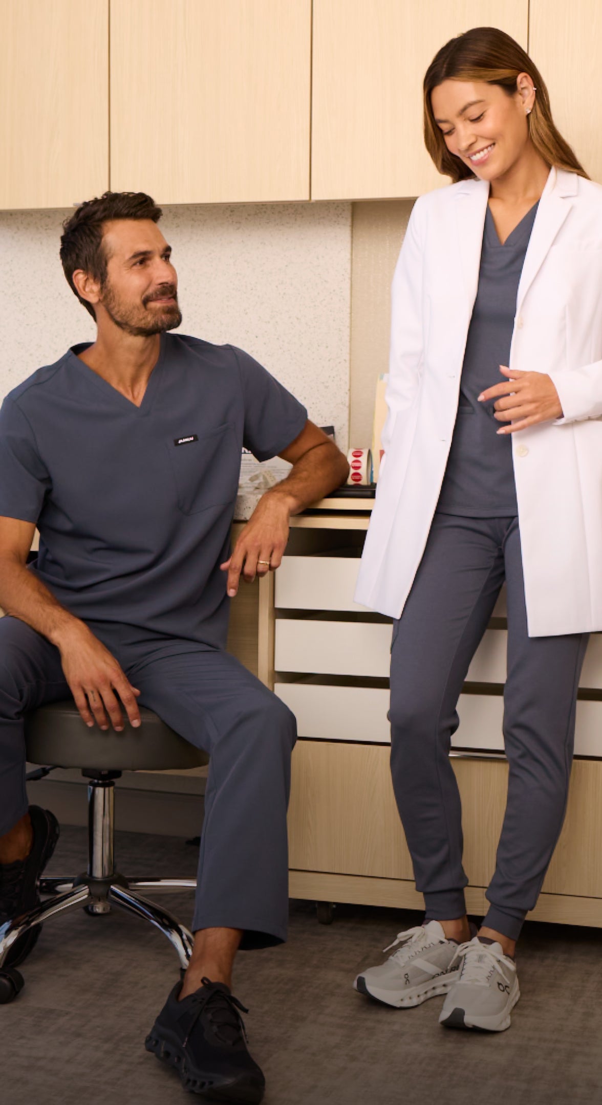 Smiling man in dark gray Jaanuu scrubs seated on a stool beside a woman in a white lab coat over dark gray scrubs in a clinic office