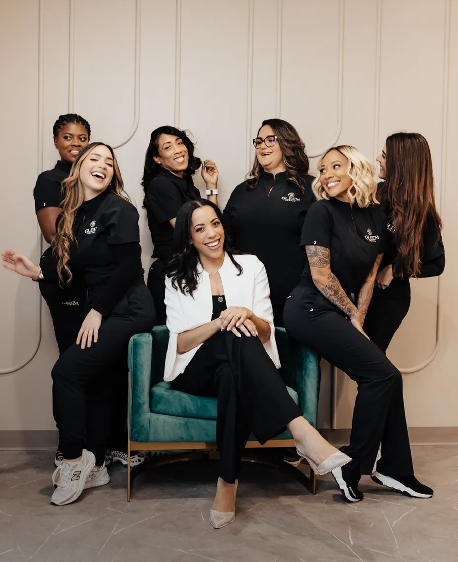 Seven women laughing together, six in black Jaanuu scrubs embroidered with the Gleem logo and one in a white blazer seated at the center in a teal velvet armchair