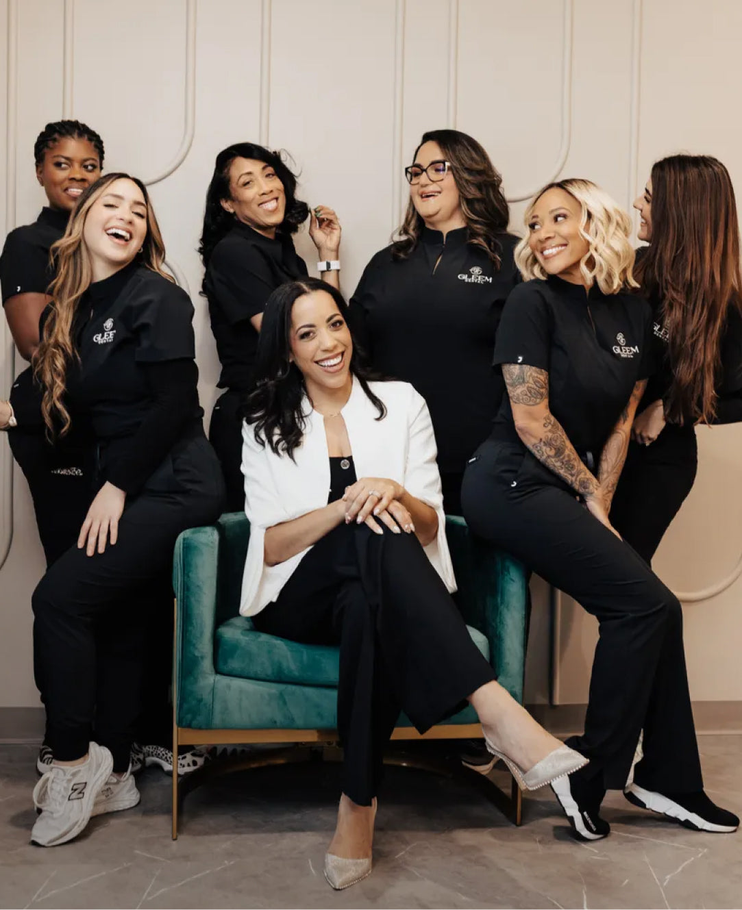 Seven women laughing together, six in black Jaanuu scrubs embroidered with the Gleem logo and one in a white blazer seated at the center in a teal velvet armchair