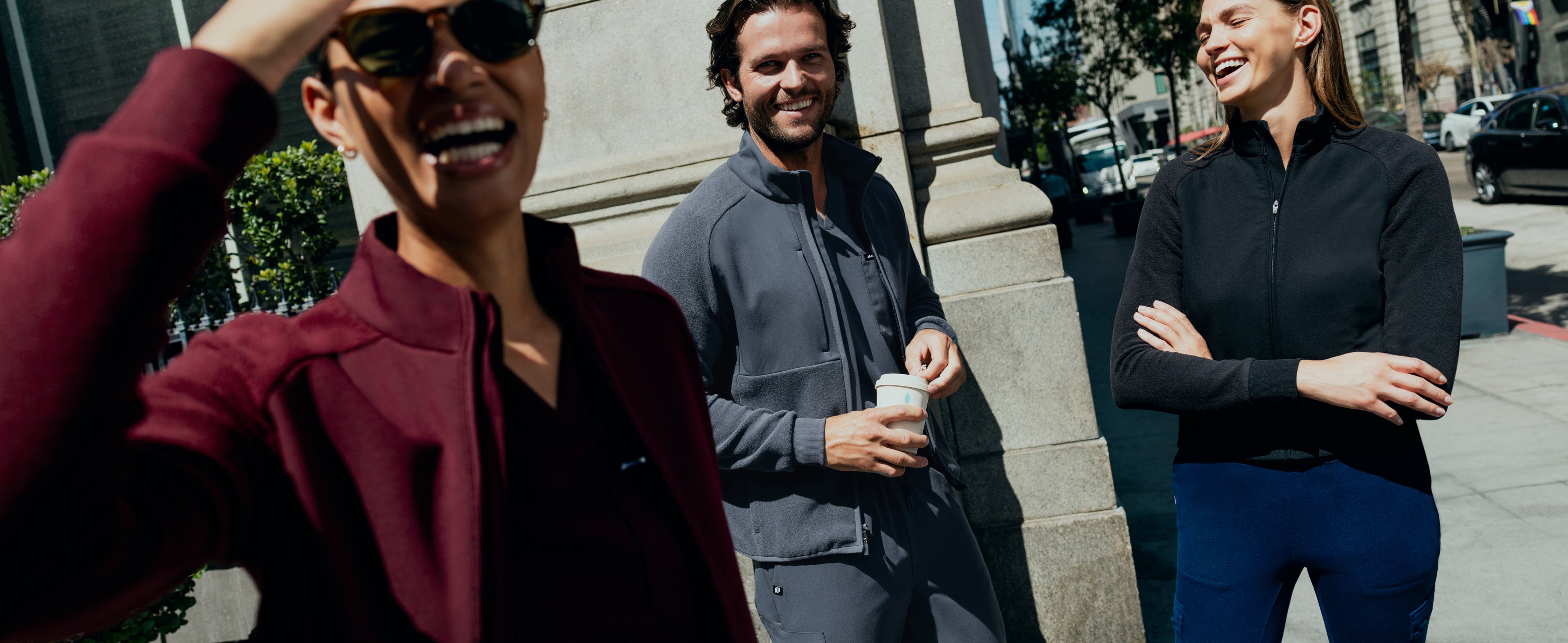 Three people in Jaanuu scrubs and zip jackets laughing on a sunny city sidewalk. A woman in sunglasses and dark red stands in front, a man in dark gray holds a coffee cup by a stone column, and a woman in a black jacket and navy blue pants crosses her arms.