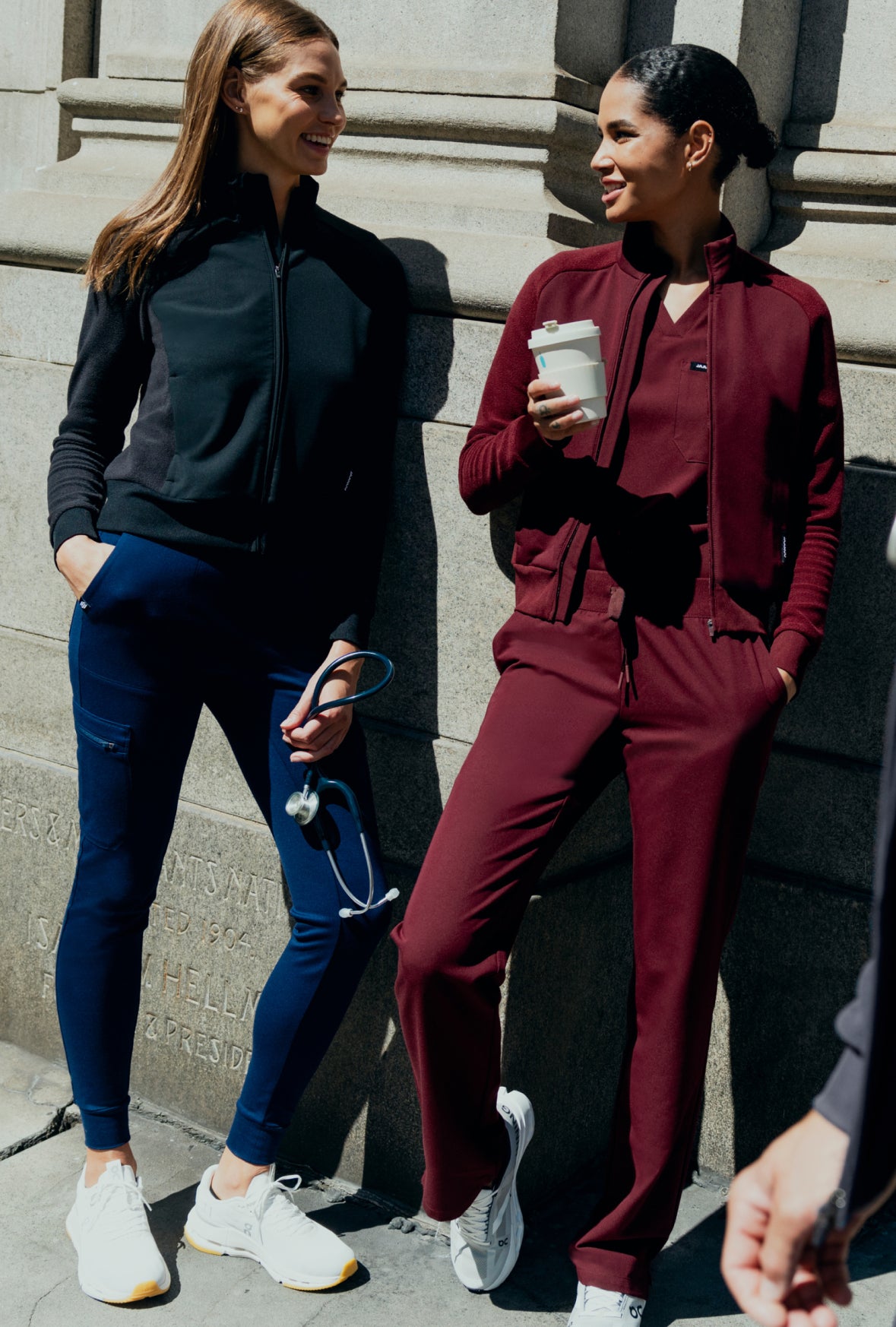 Two women in Jaanuu scrubs and zip jackets chatting against a sunlit stone building. One wears a black jacket with navy blue joggers and holds a stethoscope, the other wears dark red and holds a coffee cup.