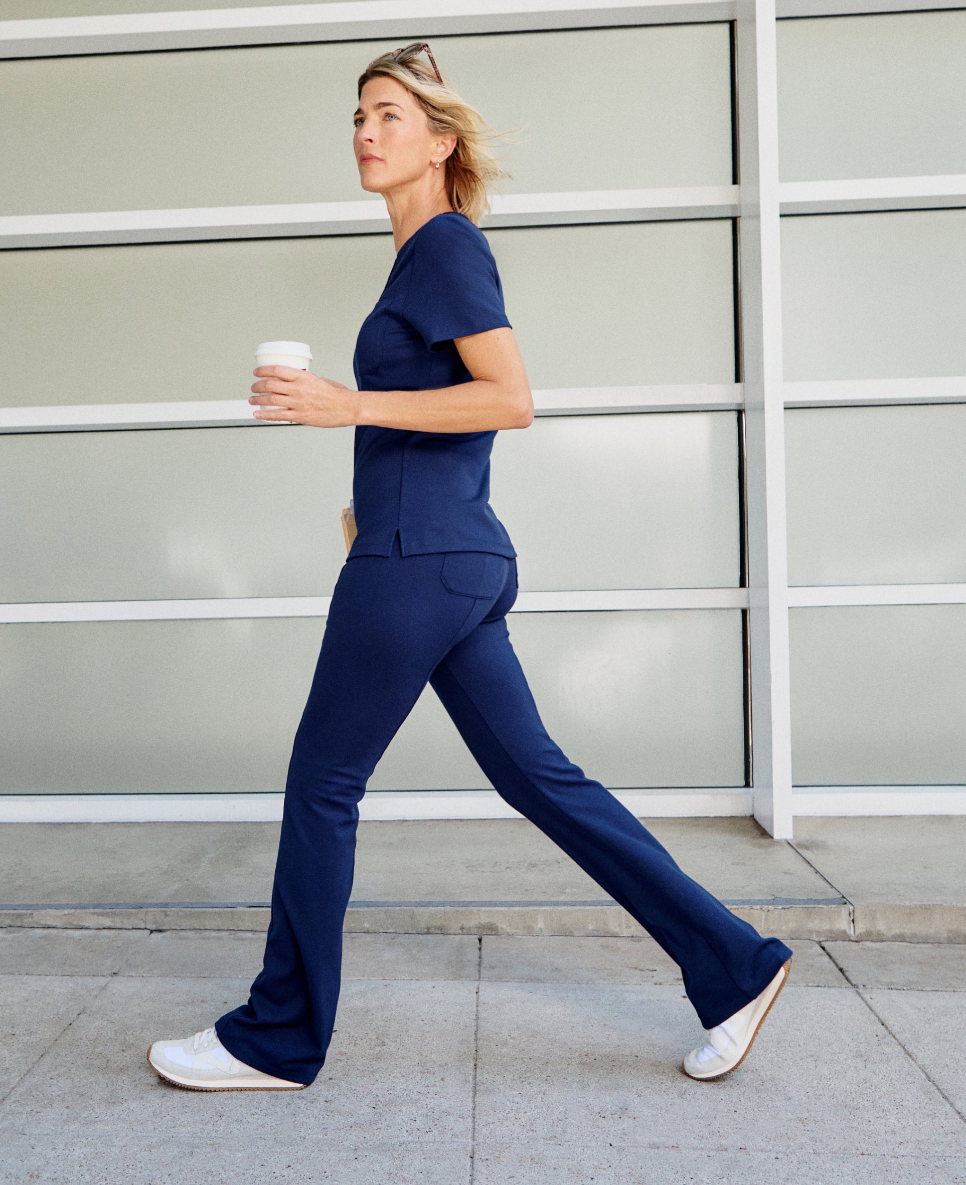 Woman in a navy Jaanuu scrub top and flared scrub pants striding past a frosted glass wall with a coffee cup in hand