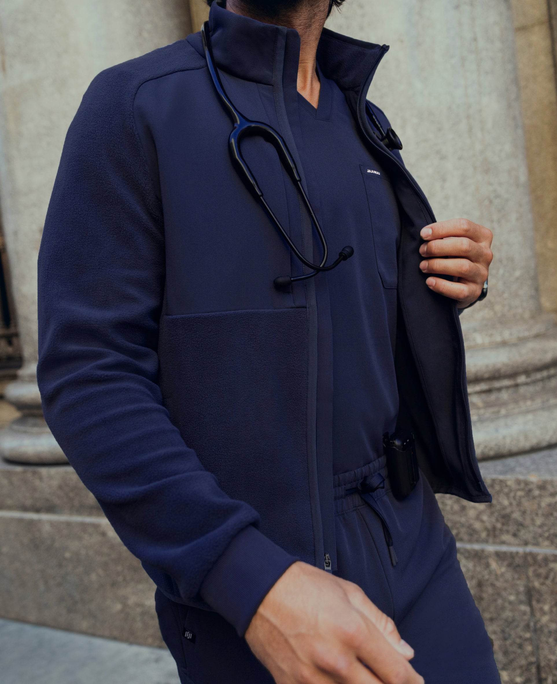 Close-up of a man in a navy Jaanuu fleece zip jacket over a navy scrub top with a stethoscope around his neck, in front of stone columns