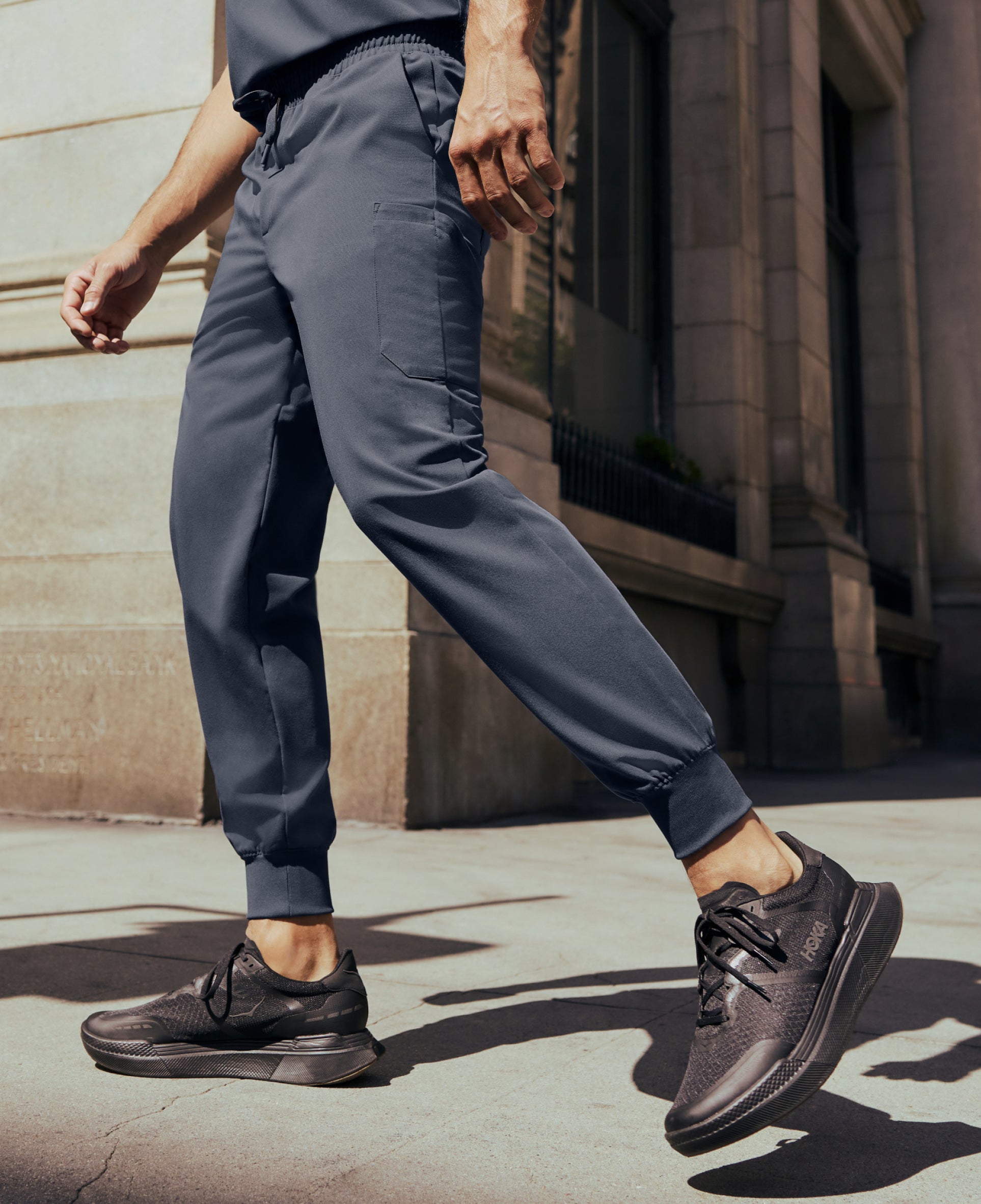 Close-up of a man's legs in charcoal gray Jaanuu jogger scrub pants and black sneakers mid-stride on a city sidewalk