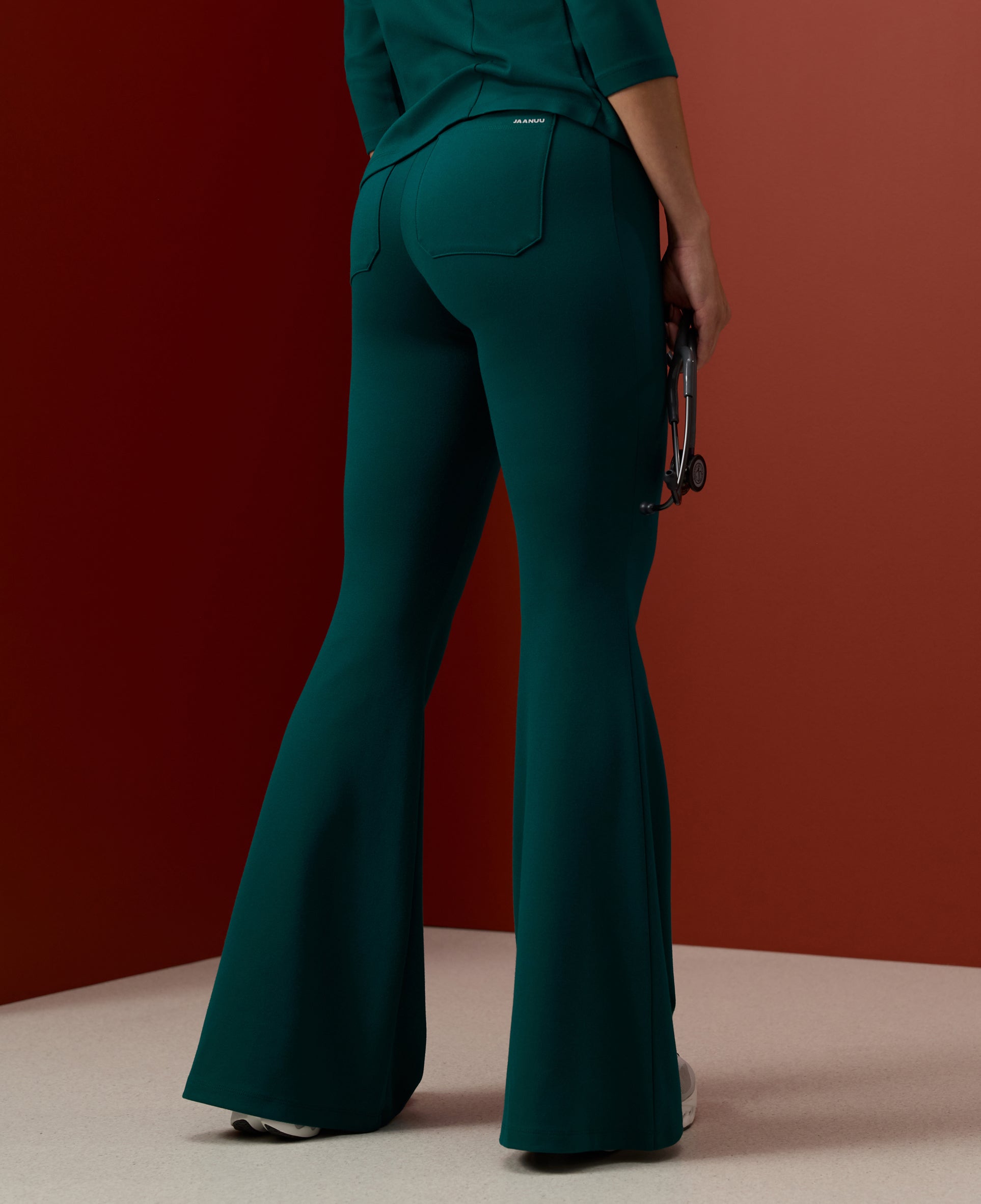Back view of a woman in dark green Jaanuu flared scrub pants and a matching top holding a stethoscope, against a rust-red backdrop