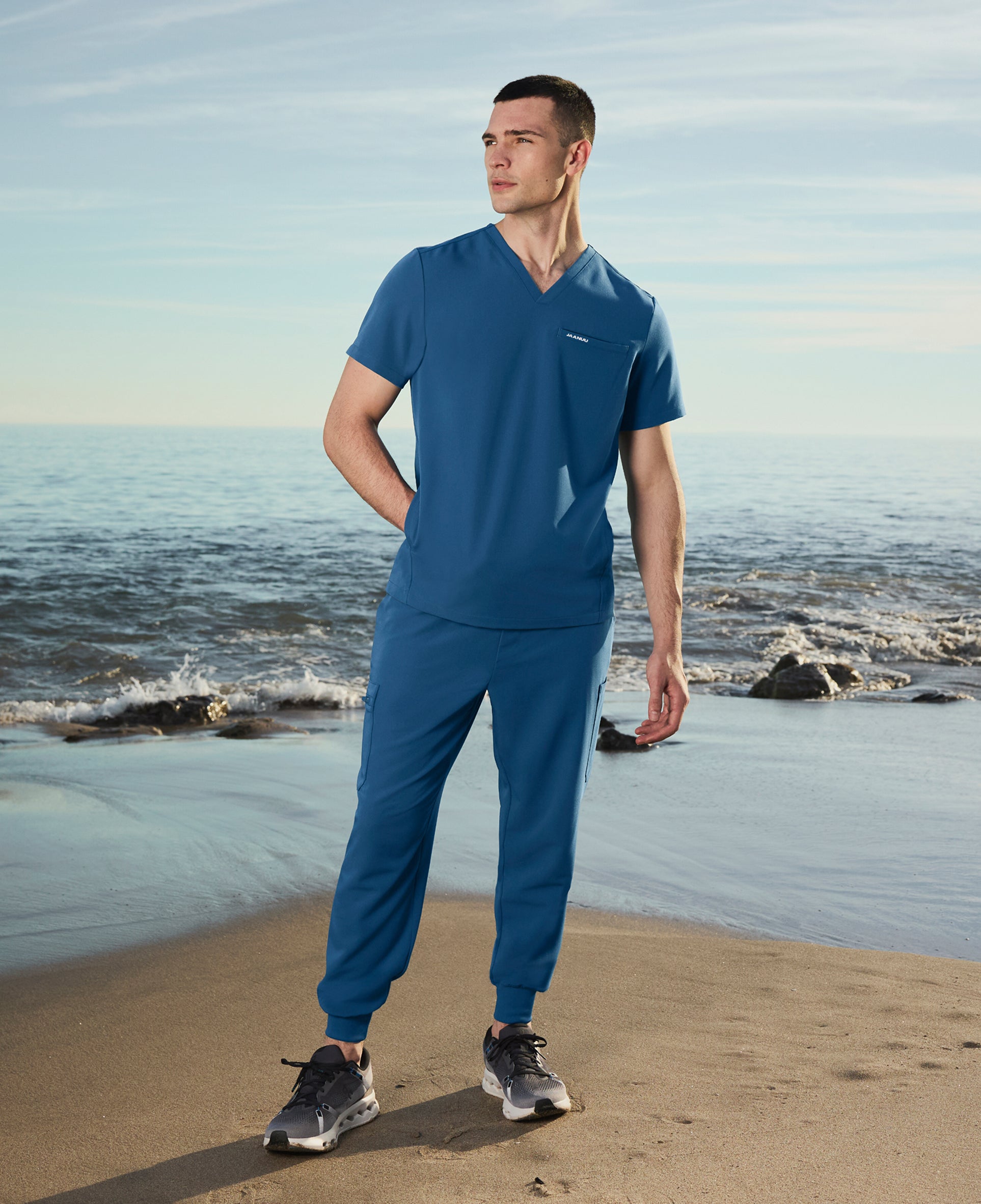 Man in a blue Jaanuu V-neck scrub top and jogger scrub pants standing on a beach with waves behind him, one hand in his pocket