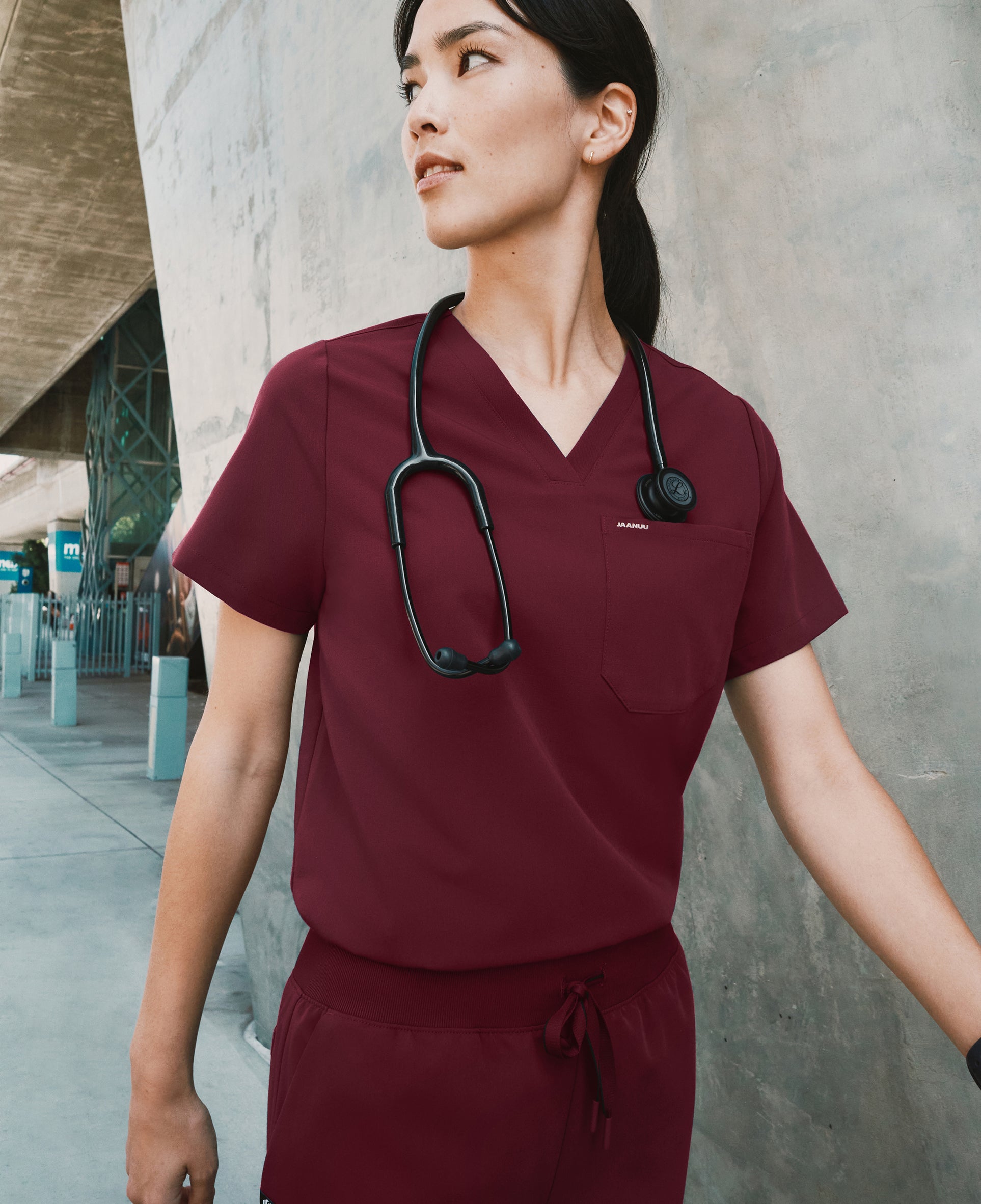 Woman in a burgundy Jaanuu V-neck scrub top and jogger scrub pants with a stethoscope around her neck walking past a concrete wall, looking over her shoulder