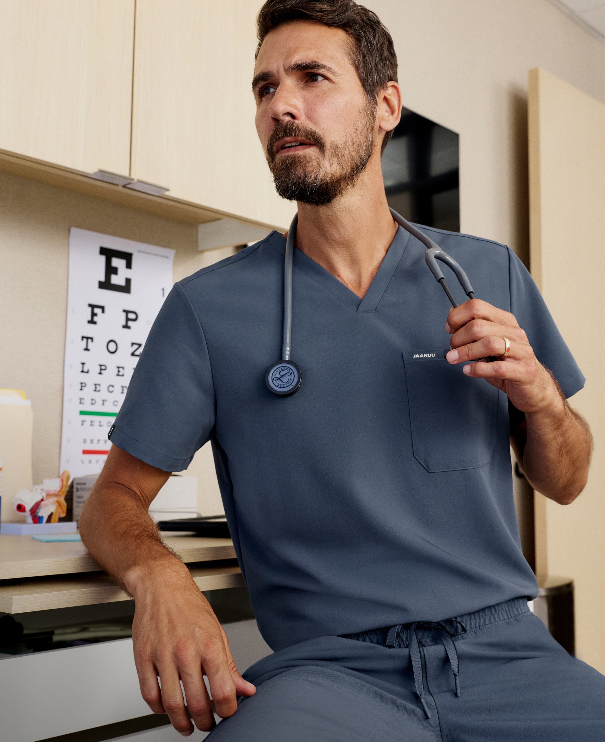 Man in a slate blue Jaanuu V-neck scrub top and scrub pants sitting at a desk in an exam room with an eye chart behind him, holding his stethoscope