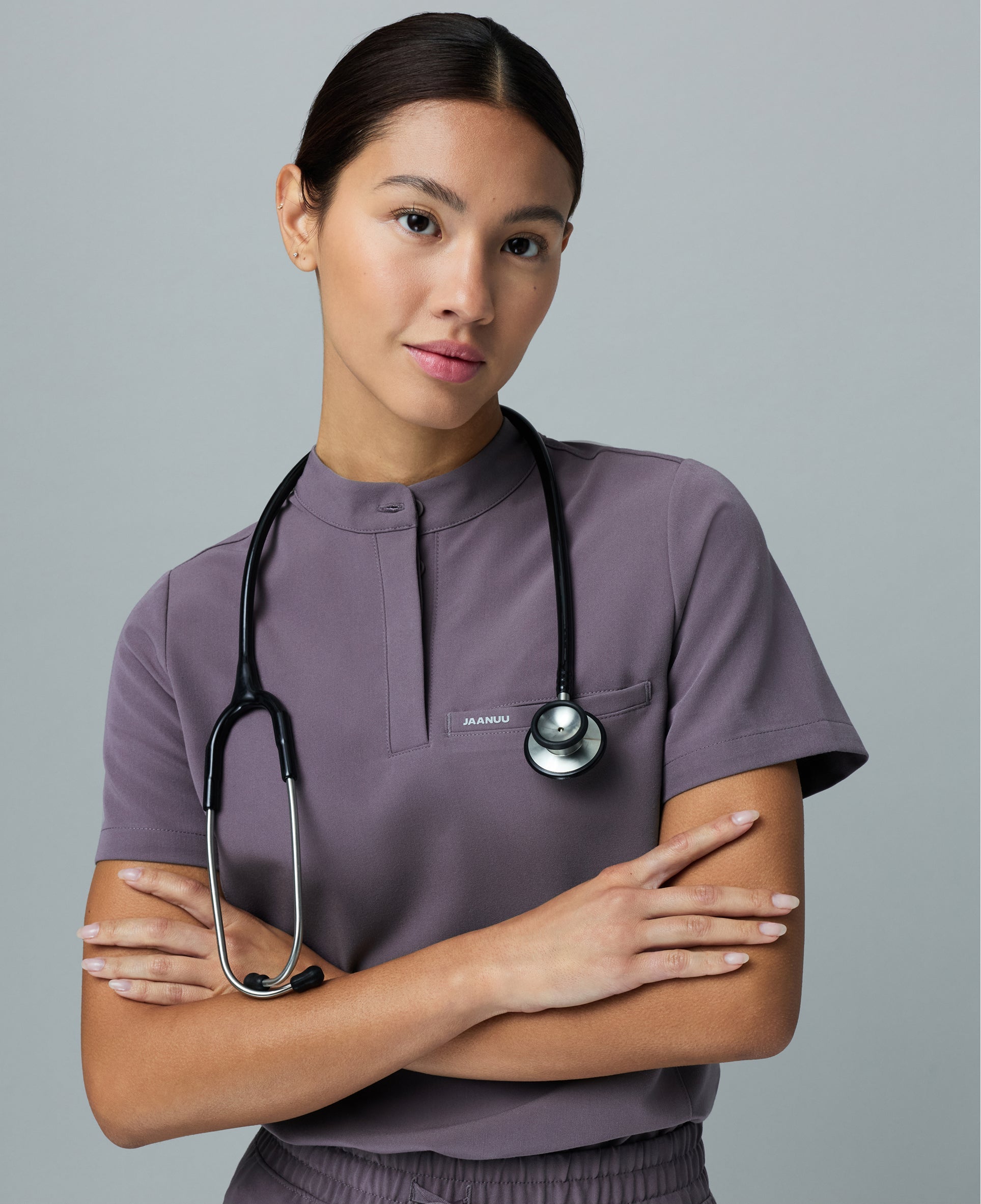 Woman in a muted purple Jaanuu stand-collar scrub top with her arms crossed and a stethoscope around her neck against a gray backdrop