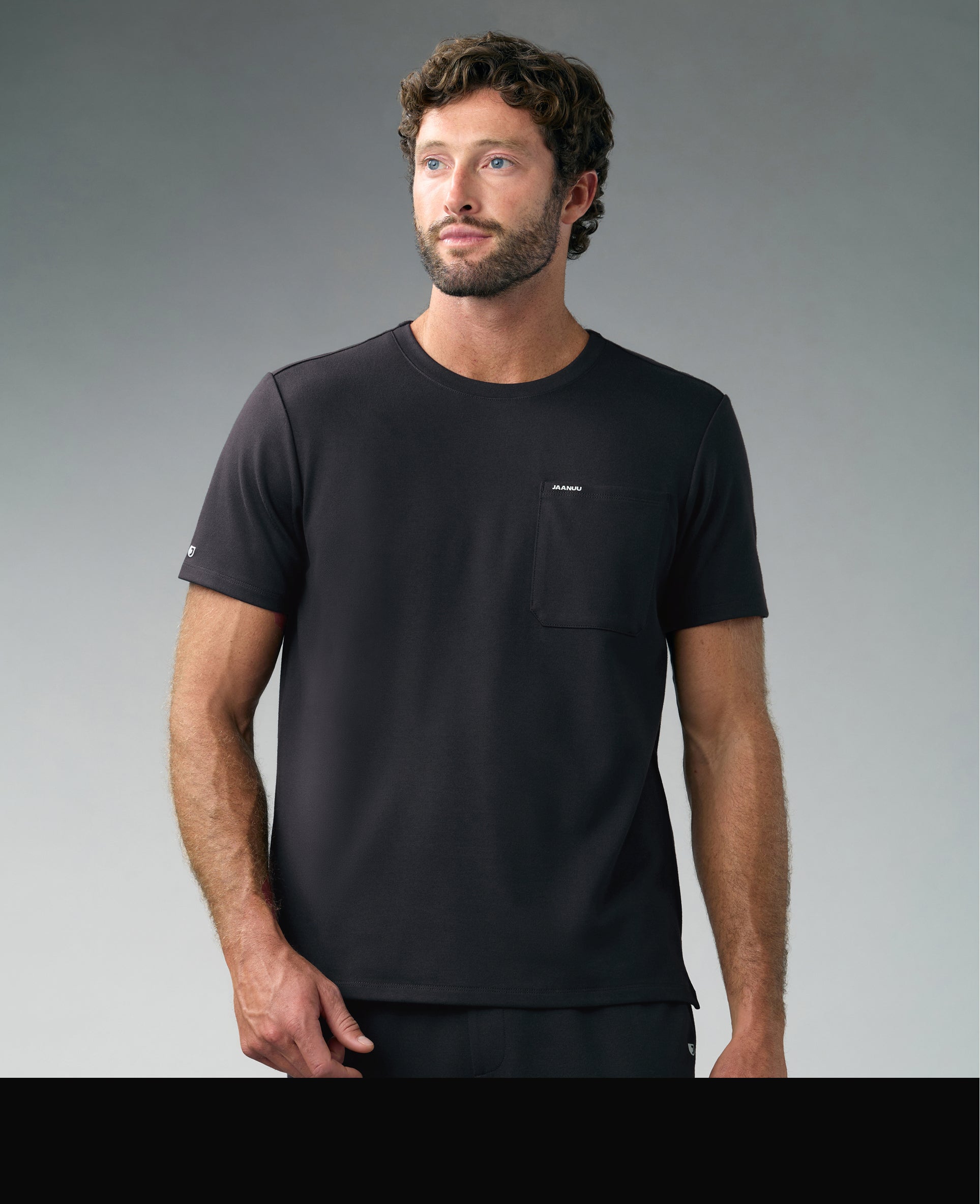 Man in a black Jaanuu crewneck pocket tee looking to the side against a gray backdrop, with a black color band across the bottom