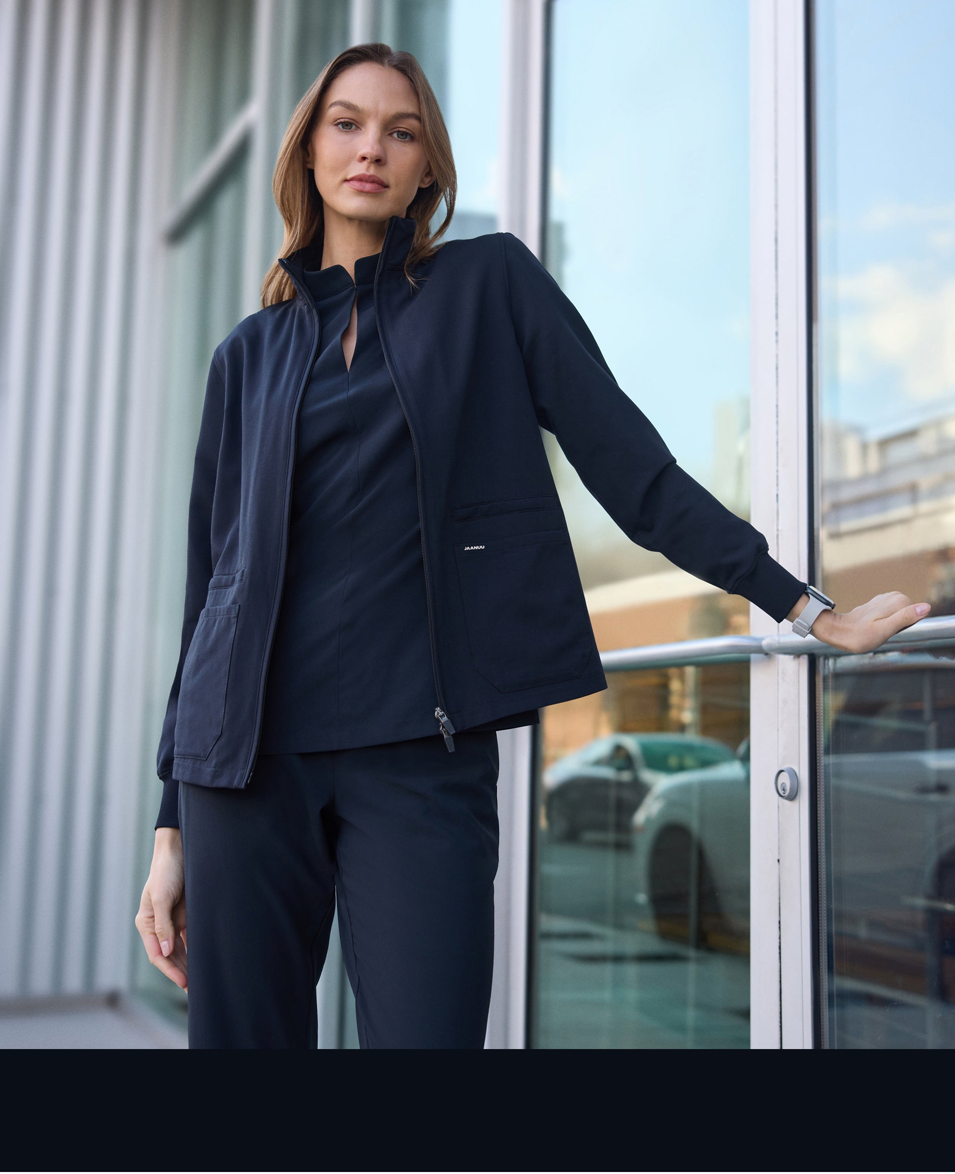Woman in a black Jaanuu zip-front scrub jacket over a black scrub top and black scrub pants, standing outside a glass building entrance with one hand on the railing, with a black color band across the bottom