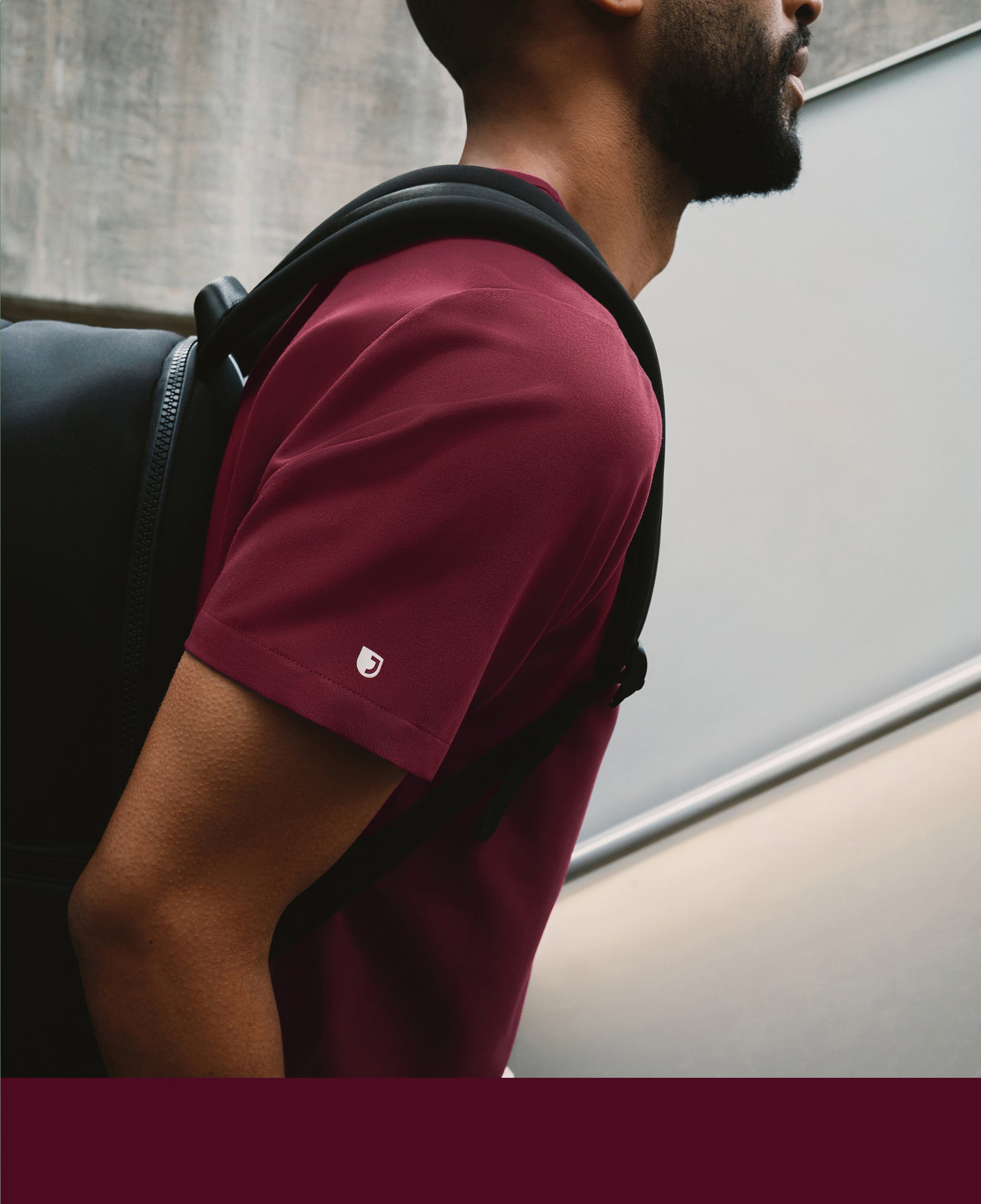 Close-up of a man in a burgundy Jaanuu scrub top with a backpack strap over his shoulder walking past a concrete wall, with a burgundy color band across the bottom