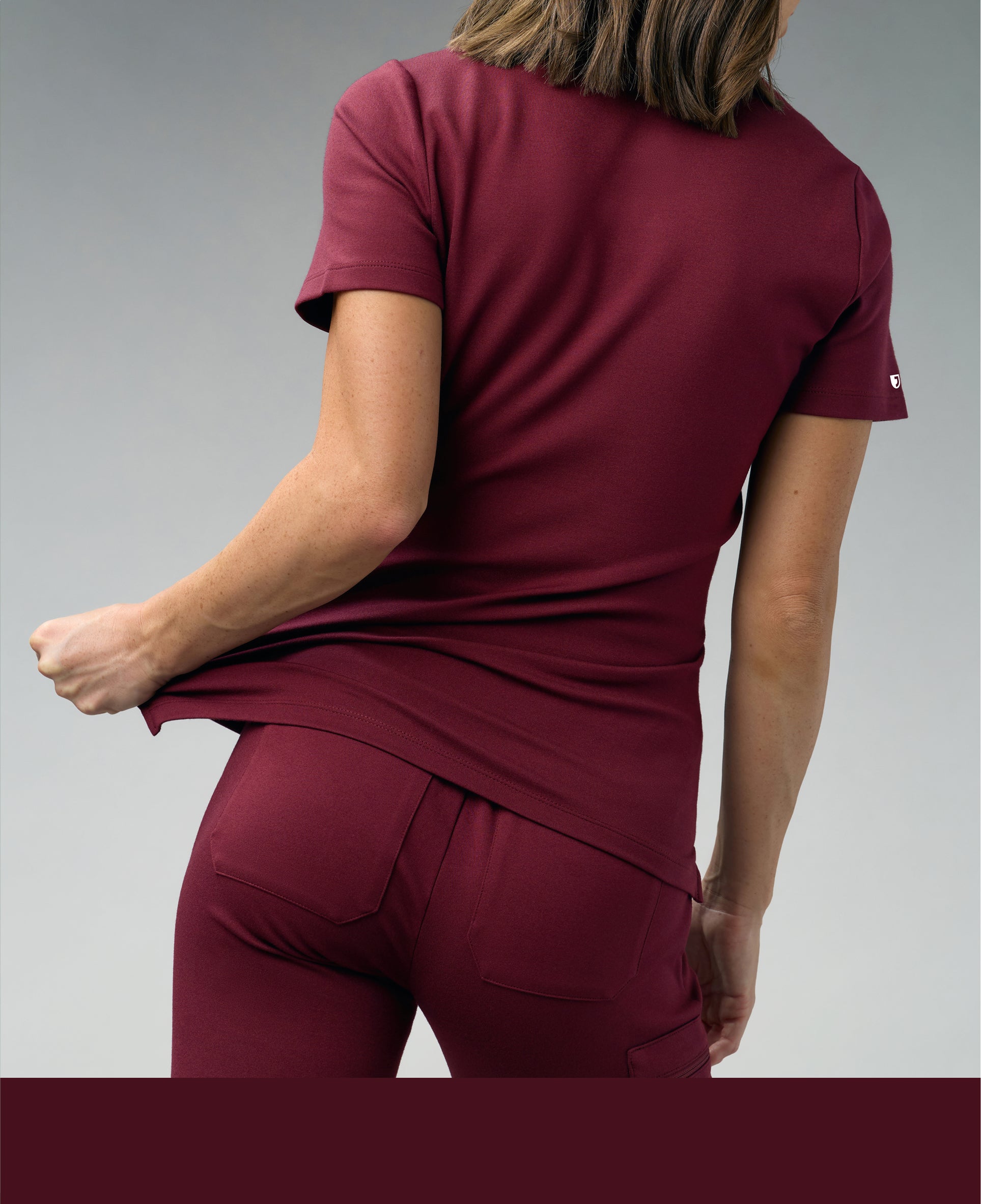 Back view of a woman in a burgundy Jaanuu scrub top and scrub pants tugging the hem of the top, against a gray backdrop, with a burgundy color band across the bottom