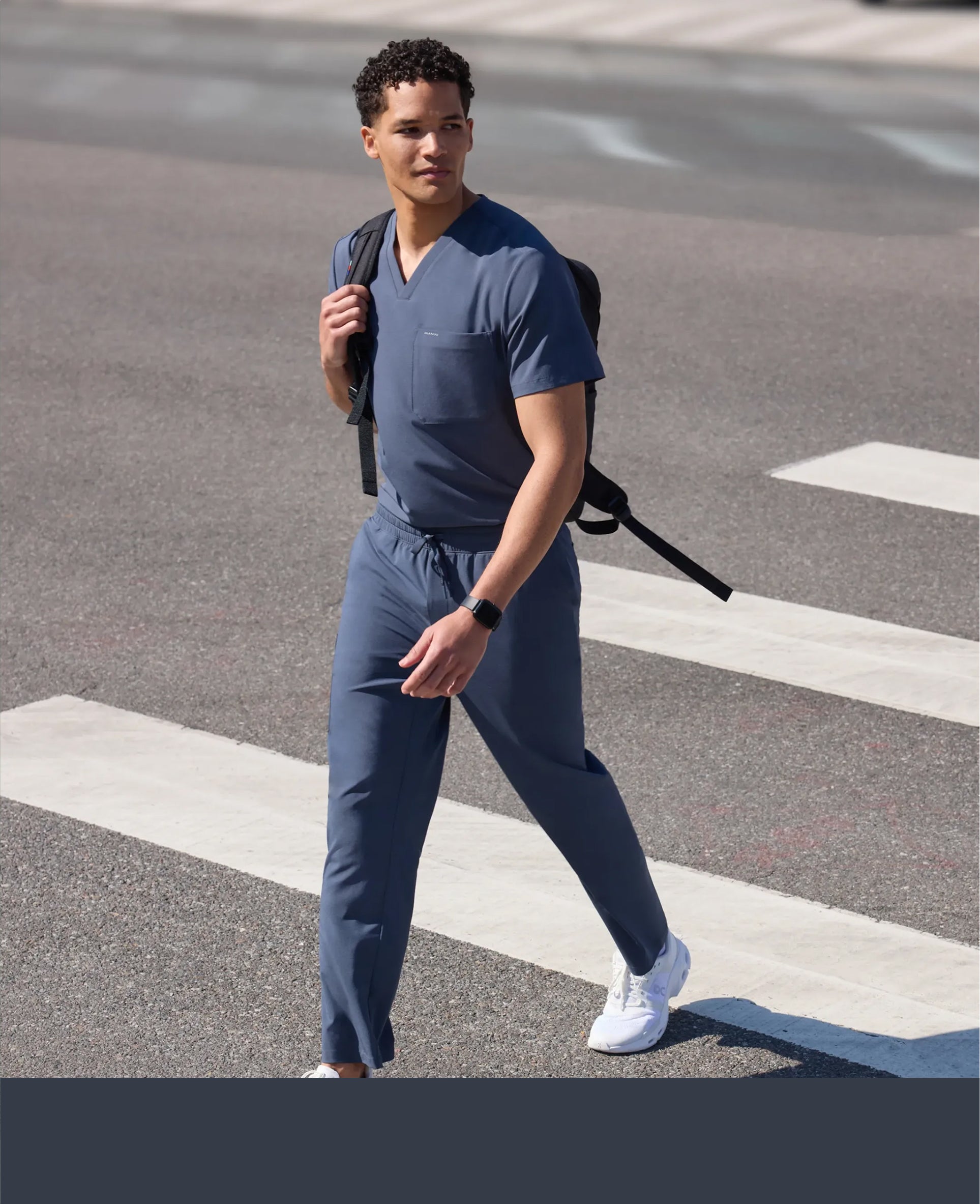 Man in a carbon gray Jaanuu V-neck scrub top and scrub pants crossing a street with a backpack over one shoulder, with a carbon gray color band across the bottom