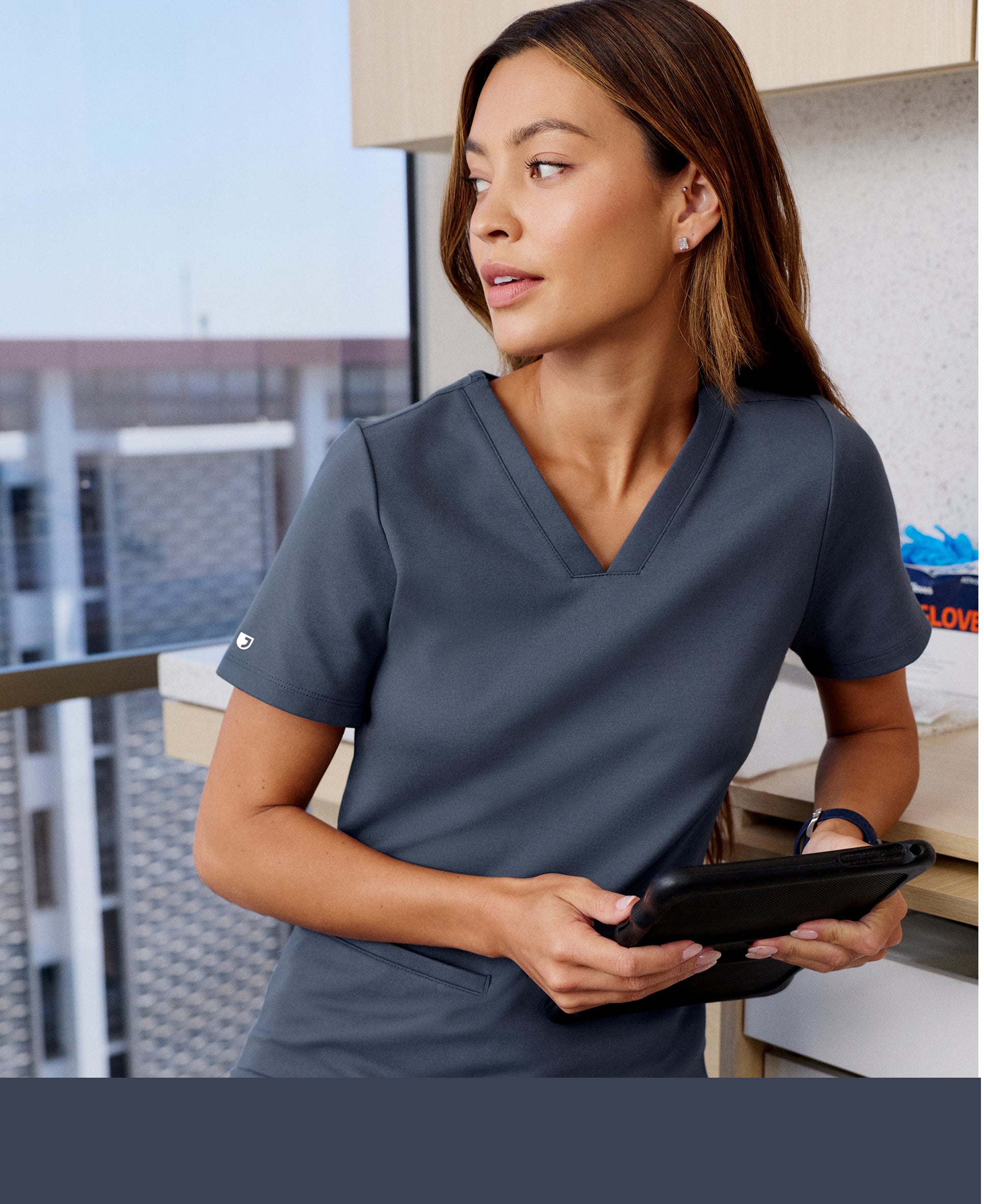 Woman in a carbon gray Jaanuu V-neck scrub top holding a tablet and looking out a clinic window, with a carbon gray color band across the bottom