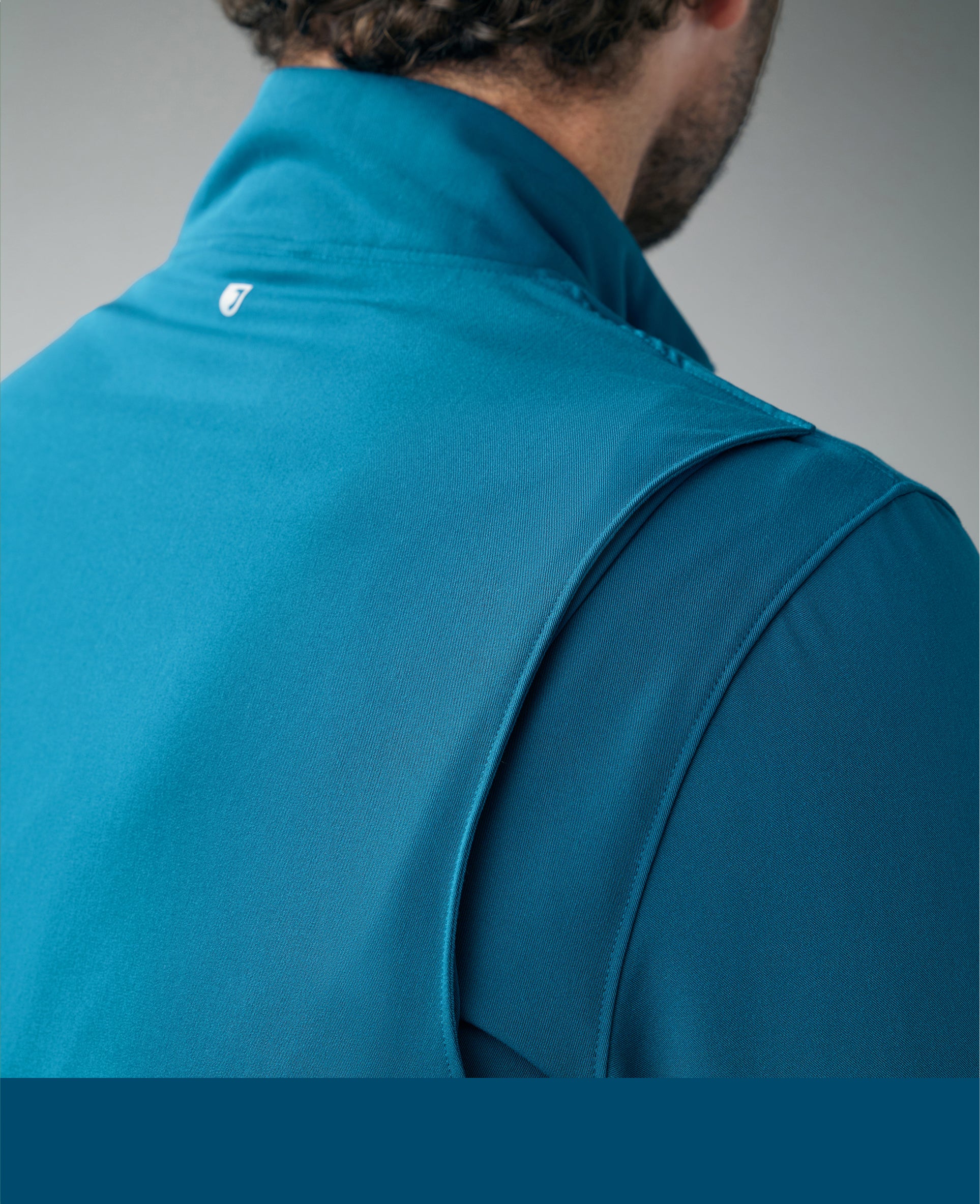 Close-up from behind of a man in a caribbean blue Jaanuu vest over a matching scrub top, with a caribbean blue color band across the bottom