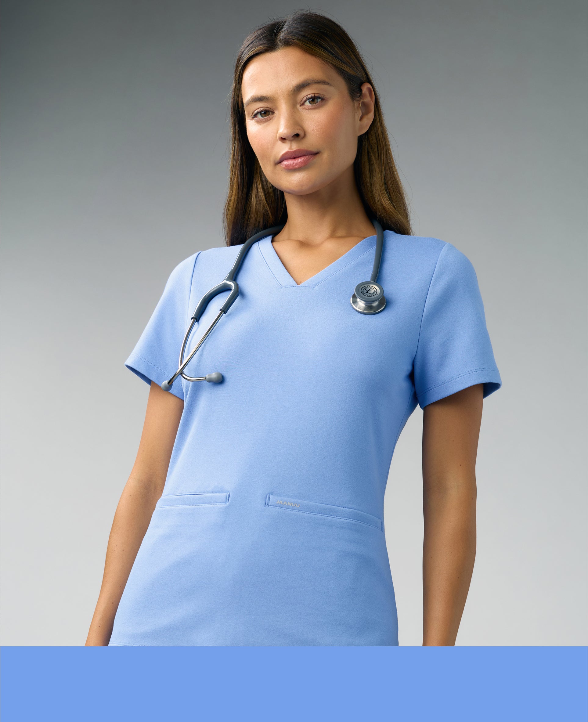 Woman in a ceil blue Jaanuu V-neck scrub top with a stethoscope around her neck facing the camera against a gray backdrop, with a ceil blue color band across the bottom