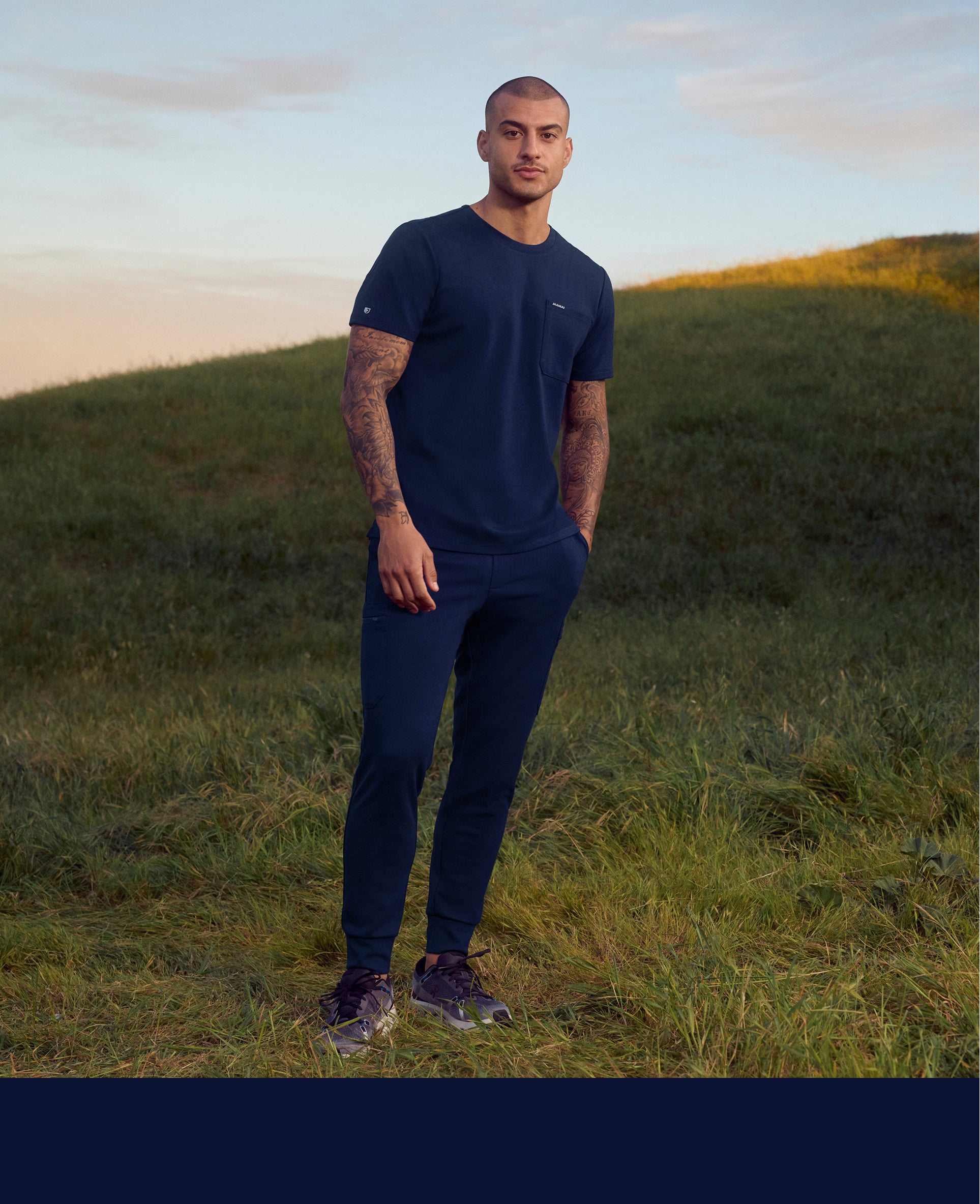 Man in a navy Jaanuu crewneck pocket tee and jogger scrub pants standing on a grassy hillside at sunset, with a navy color band across the bottom