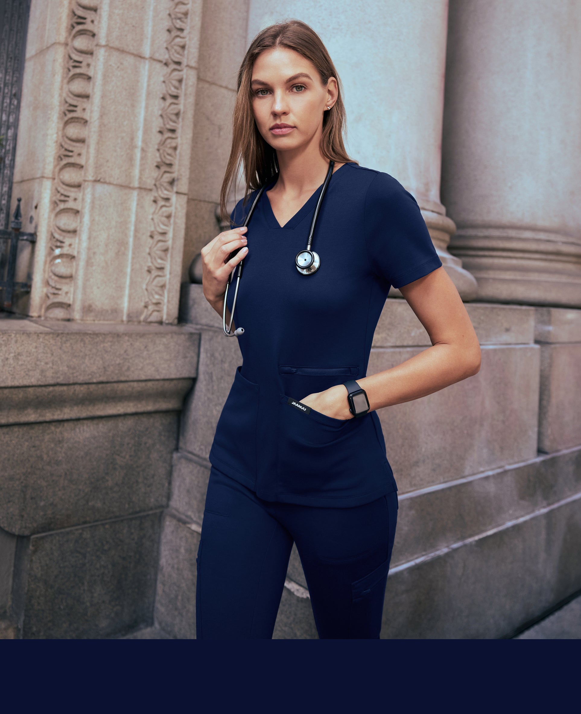 Woman in a navy Jaanuu V-neck scrub top and cargo scrub pants with a stethoscope around her neck and one hand in her pocket, standing between stone columns, with a navy color band across the bottom