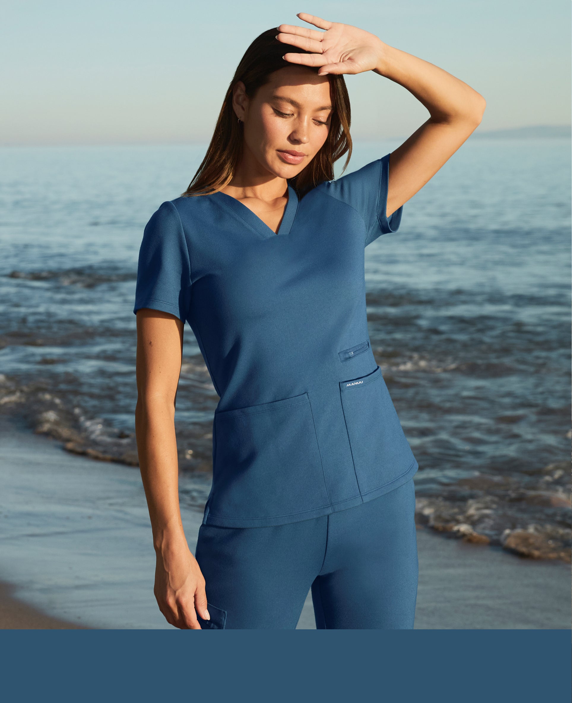 Woman in an ocean blue Jaanuu V-neck scrub top and cargo scrub pants shading her eyes at the shoreline with waves behind her, with an ocean blue color band across the bottom